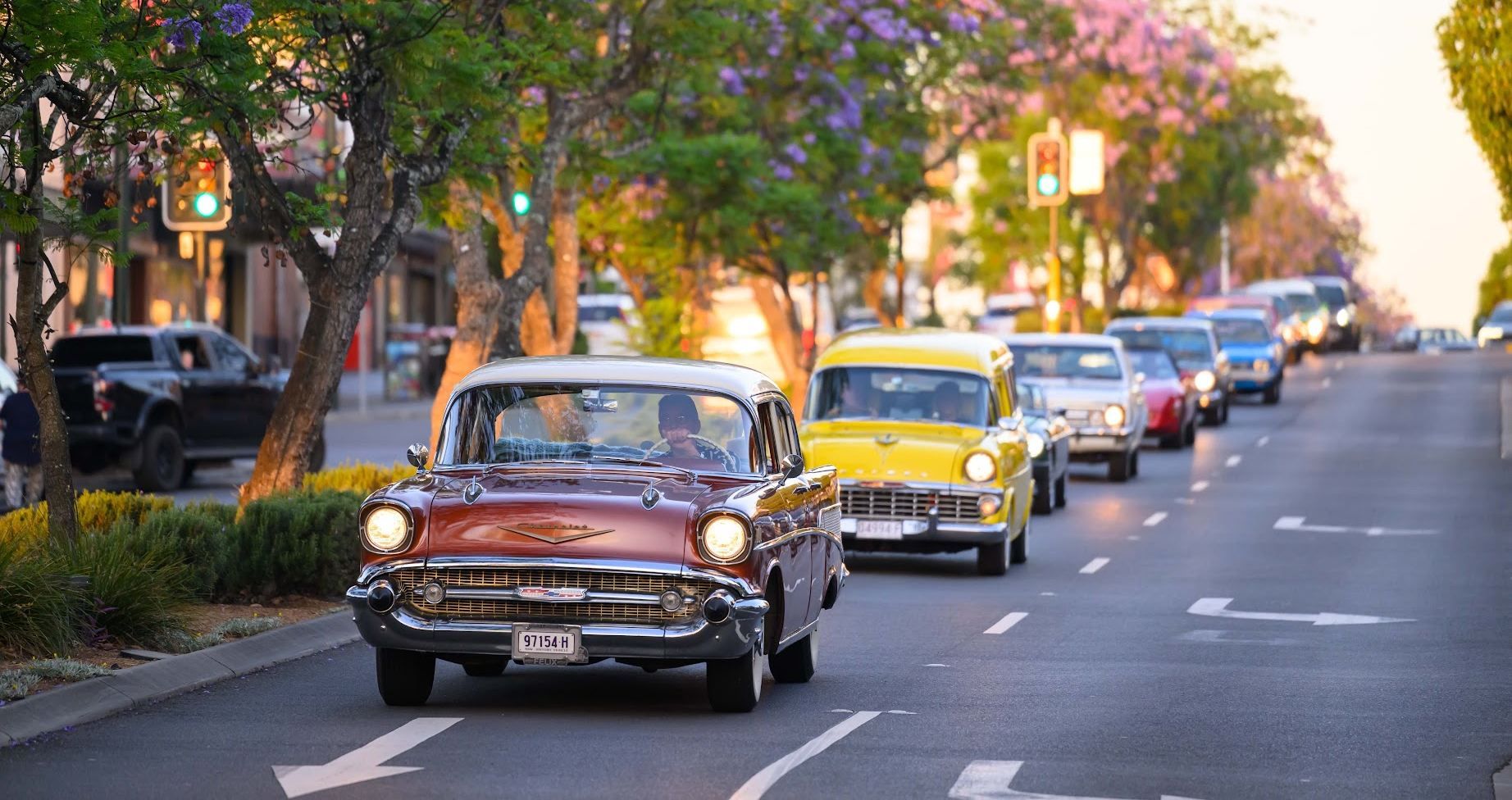 Vintage cars in a colorful parade, led by a maroon car, on a tree-lined street with blooming flowers and traffic lights.