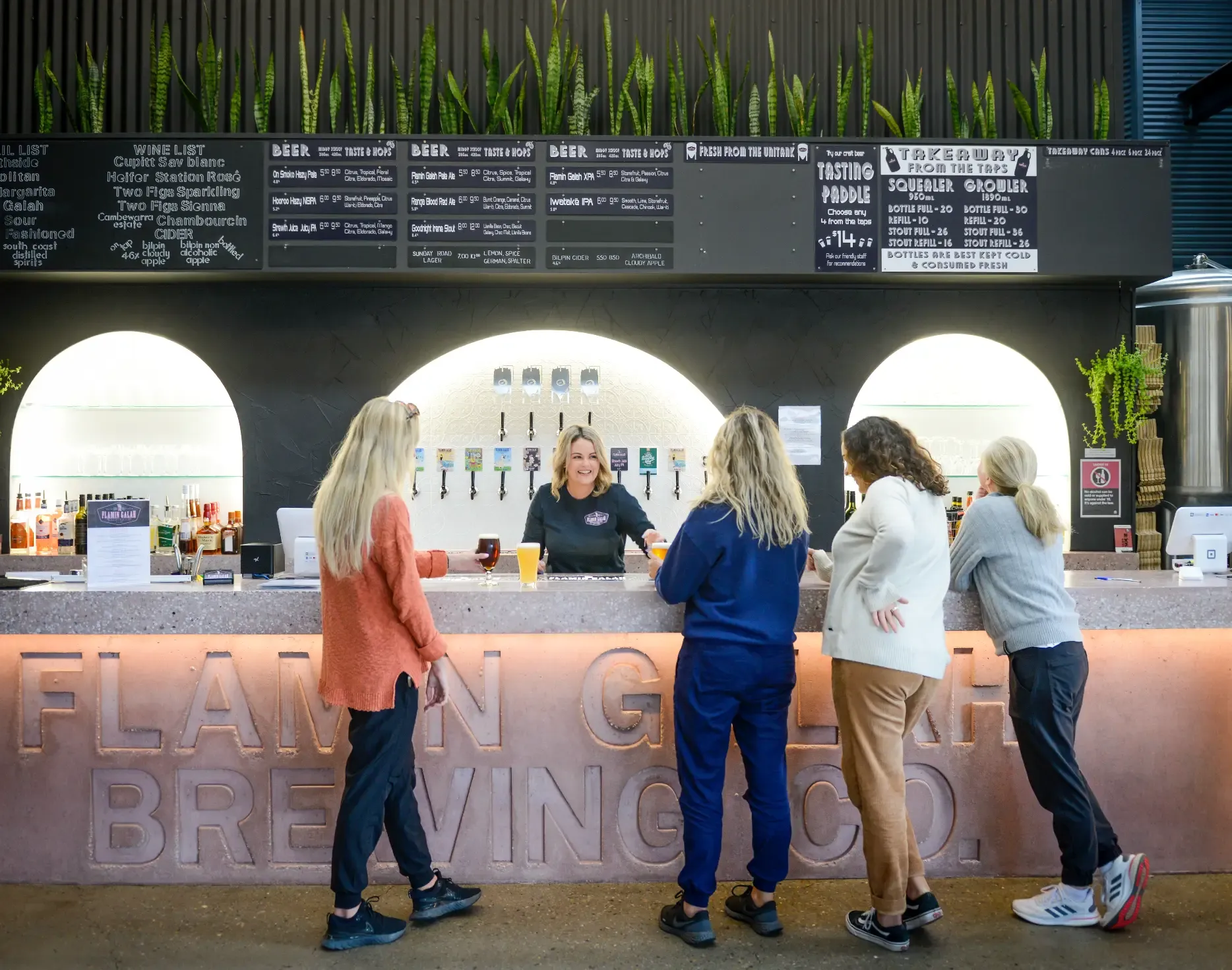 A group of women stand at a brewery bar, engaged in conversation. Beer taps and menu boards are visible, with plants above the bar.