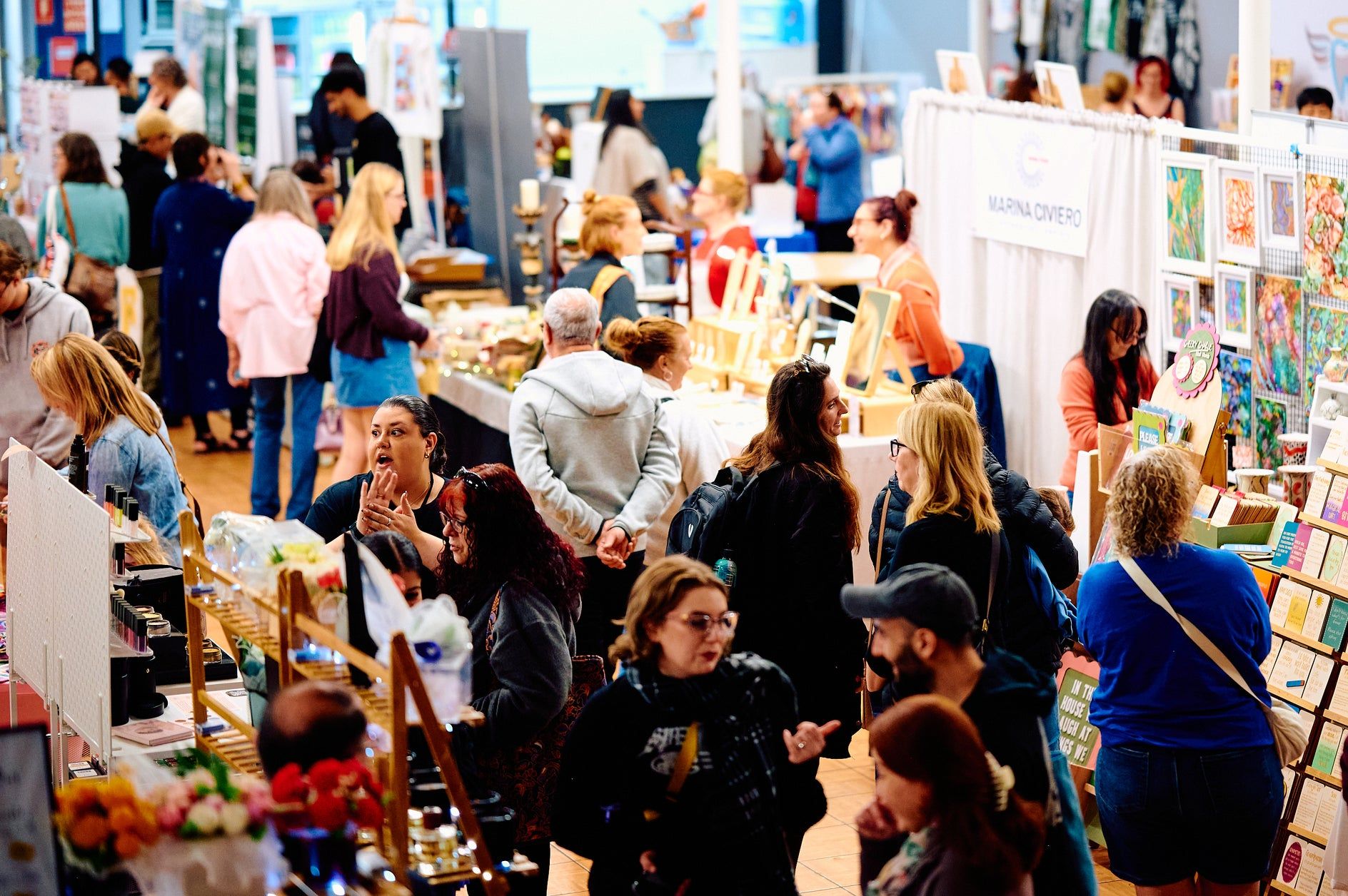 People browse various stalls at a bustling indoor market or fair, filled with colorful art, crafts, and products on display.