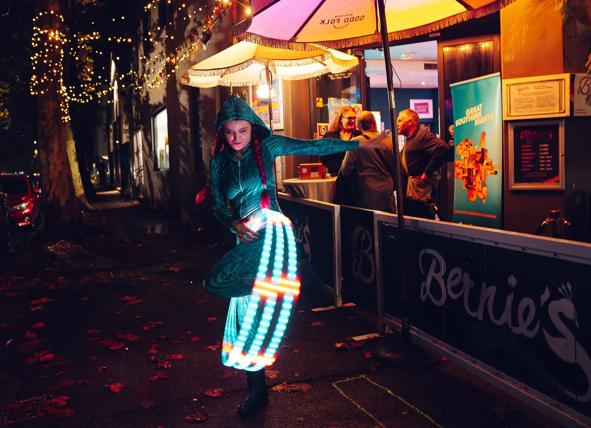 A person dances in a glowing LED outfit on a dimly lit street near a food stand, with string lights and autumn leaves scattered on the ground.