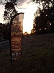 A flag advertising burgers with text and a phone number stands on a grassy roadside, silhouetted against a sunset with trees in the background.