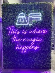 Purple neon sign on a leafy green wall reading "This is where the magic happens" with a stylized "AF" logo above.