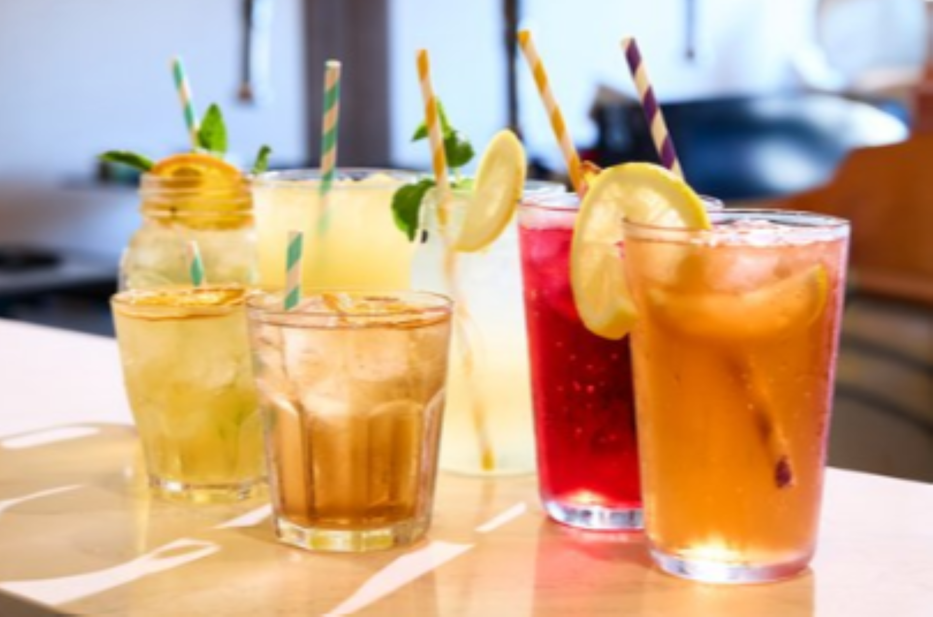 A variety of colorful drinks with straws and garnishes, including lemon slices and mint, served in different glasses on a light-colored surface.