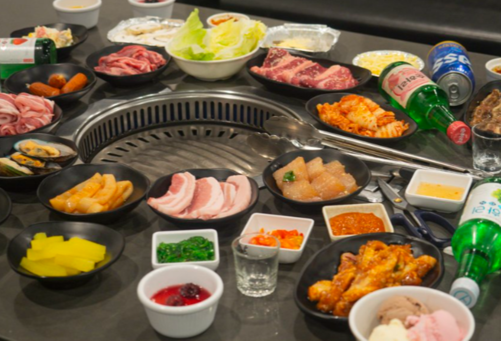 A Korean BBQ setup with assorted meats, vegetables, sauces, and drinks arranged around a grill on a dining table.
