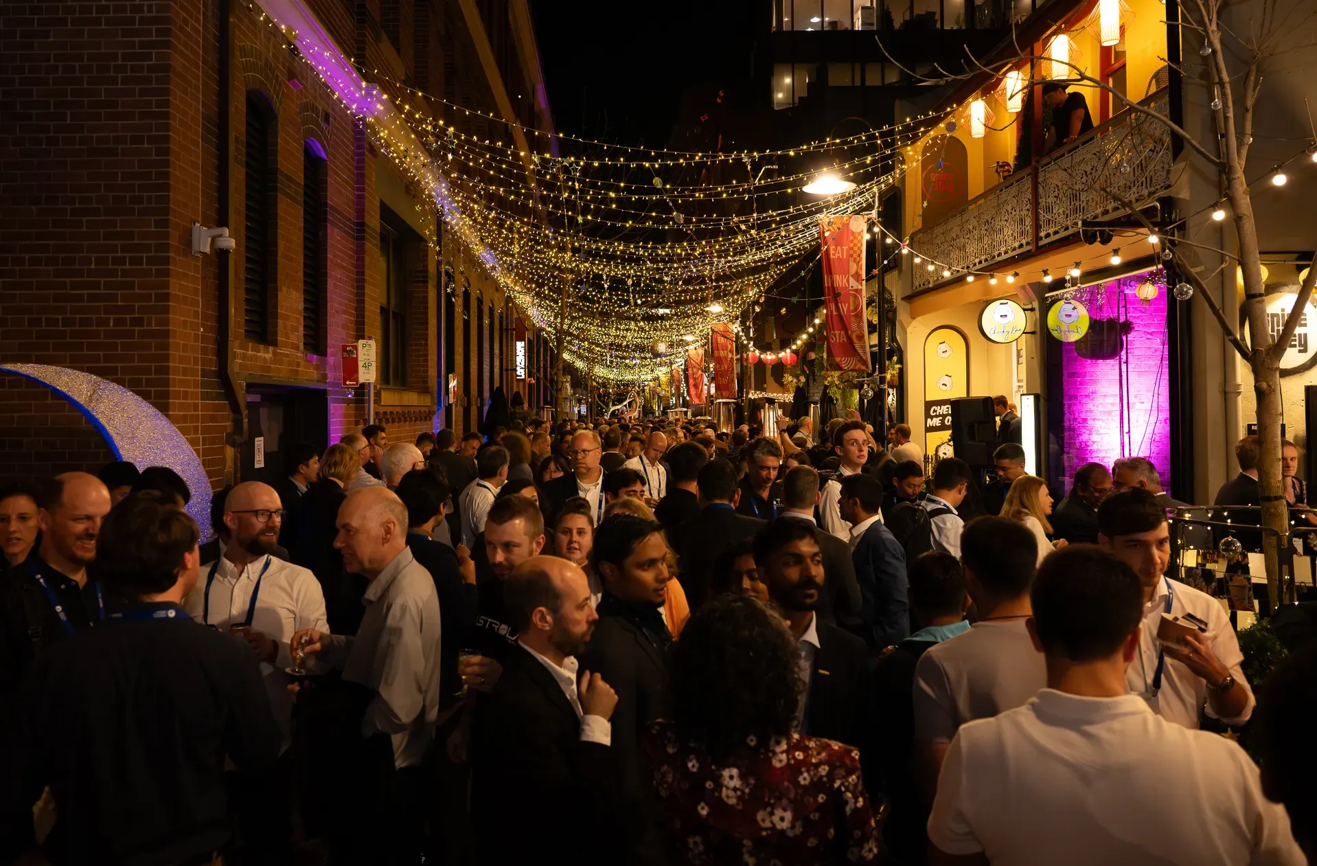 A bustling nighttime street event with a crowd of people under strings of decorative lights, surrounded by brick buildings and colorful signs.