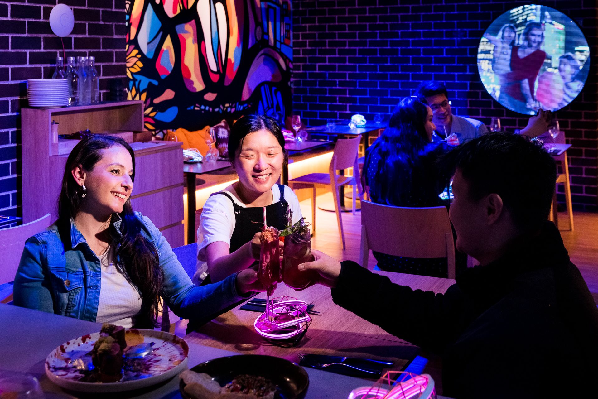 A small group of diners clinking glasses at Auvers Café in Darling Harbour with colourful ambient lighting during Vivid Sydney.