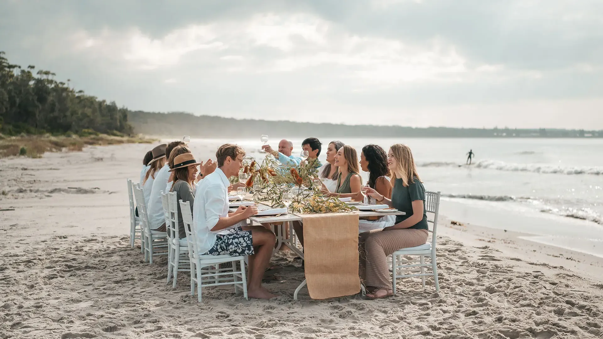 A group of people sitting at a decorated table on a sandy beach, enjoying a meal with drinks, under a cloudy sky.