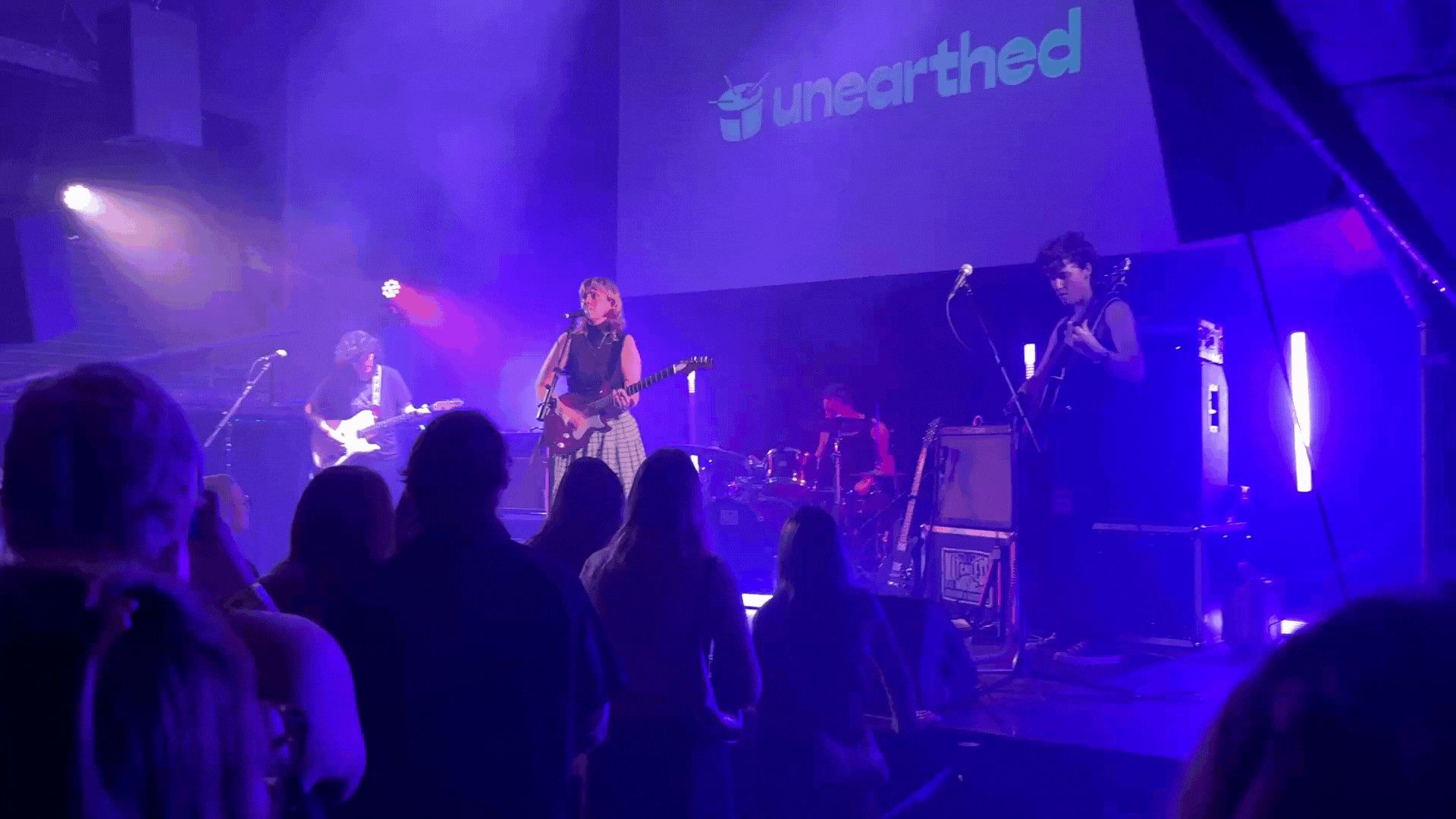 A band performs on stage with colorful lighting and a "unearthed" banner. Audience members watch from the foreground.