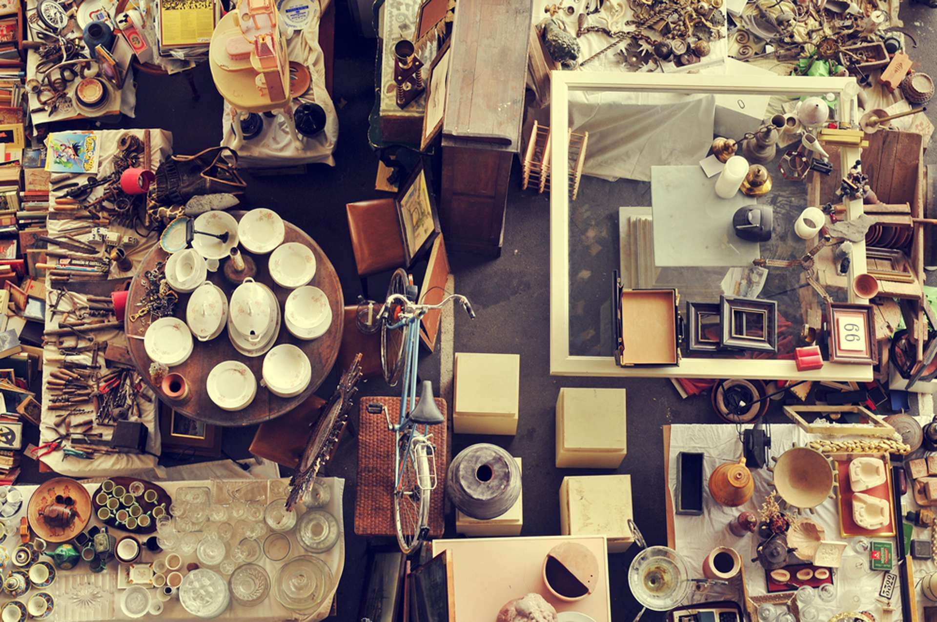 Aerial view of a cluttered flea market stall displaying dishes, frames, books, and various miscellaneous items on tables and the floor.
