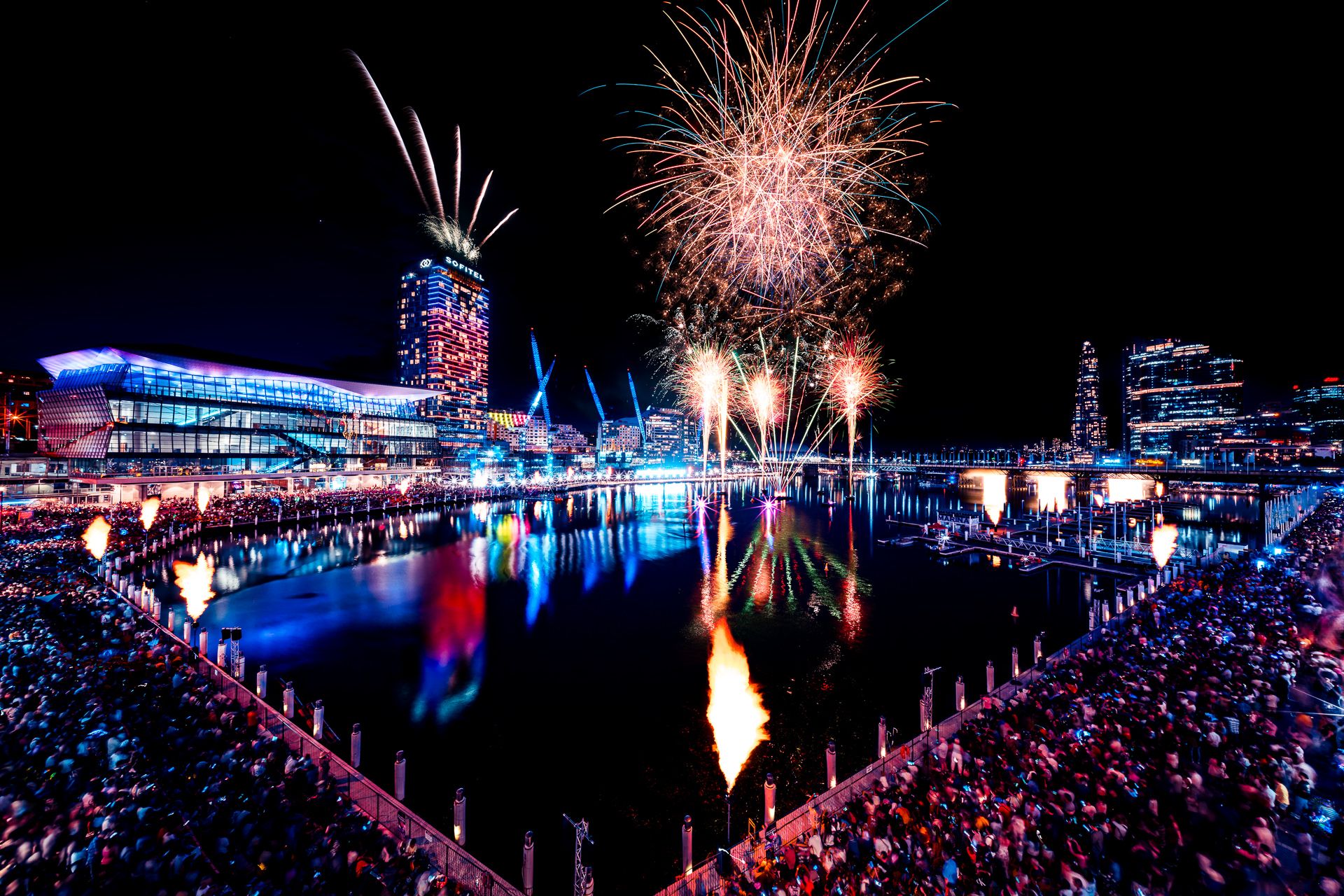 New Year’s Eve fireworks over Darling Harbour, with crowds gathered along the waterfront and the ICC Sydney and Sofitel hotel illuminated in the background.