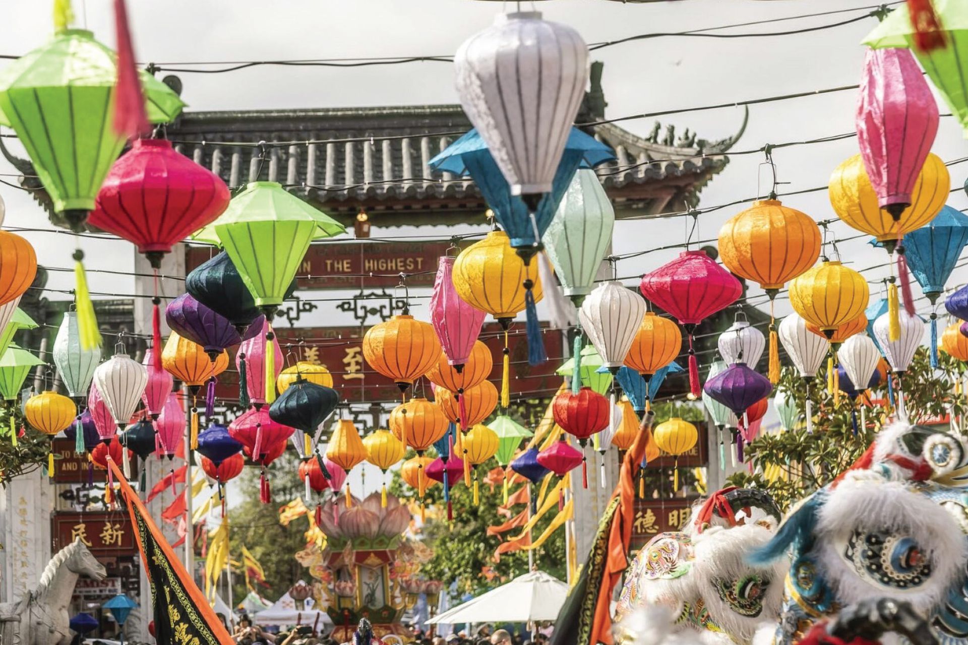 Colourful lanterns hang above a bustling street during a festival, with a traditional archway and dragon dance in the background.
