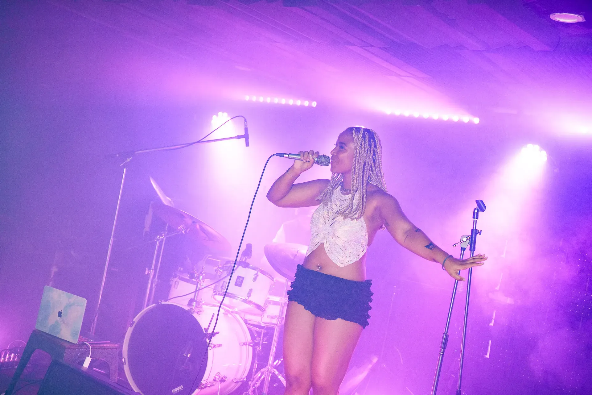 A woman with braided hair sings passionately on stage, holding a microphone under purple stage lights, with a drum set behind her.