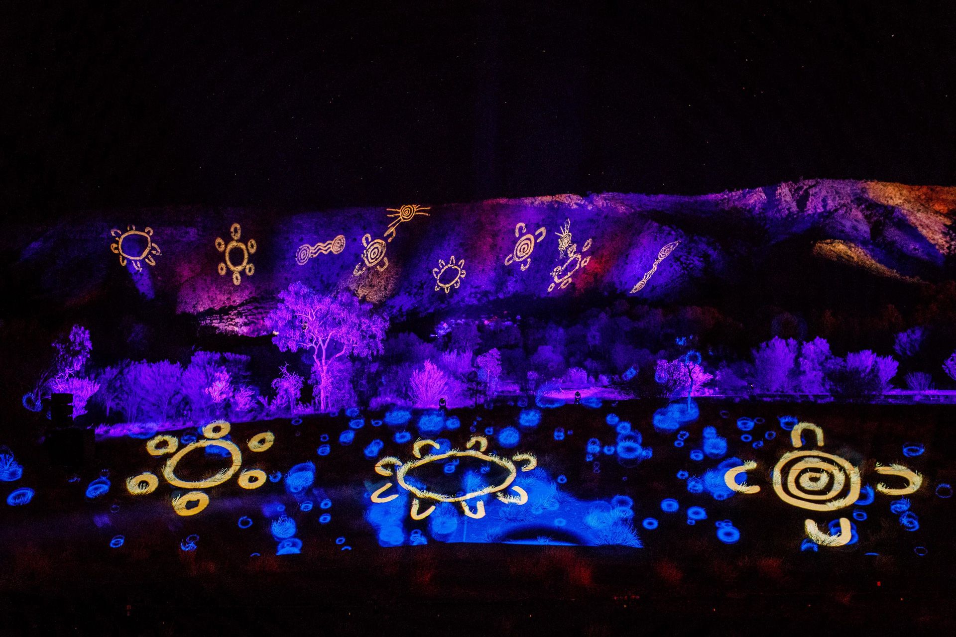 A mountain range and foreground illuminated in saturated colours and populated with First Nations symbols