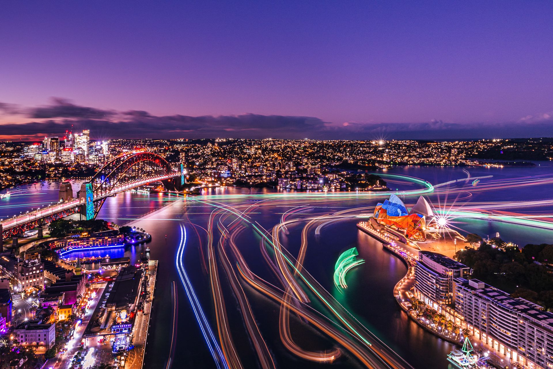 A wide sunset view over Sydney Harbour featuring the colourful projections on the Sydney Harbour Bridge and Sydney Opera House during Vivid Sydney.