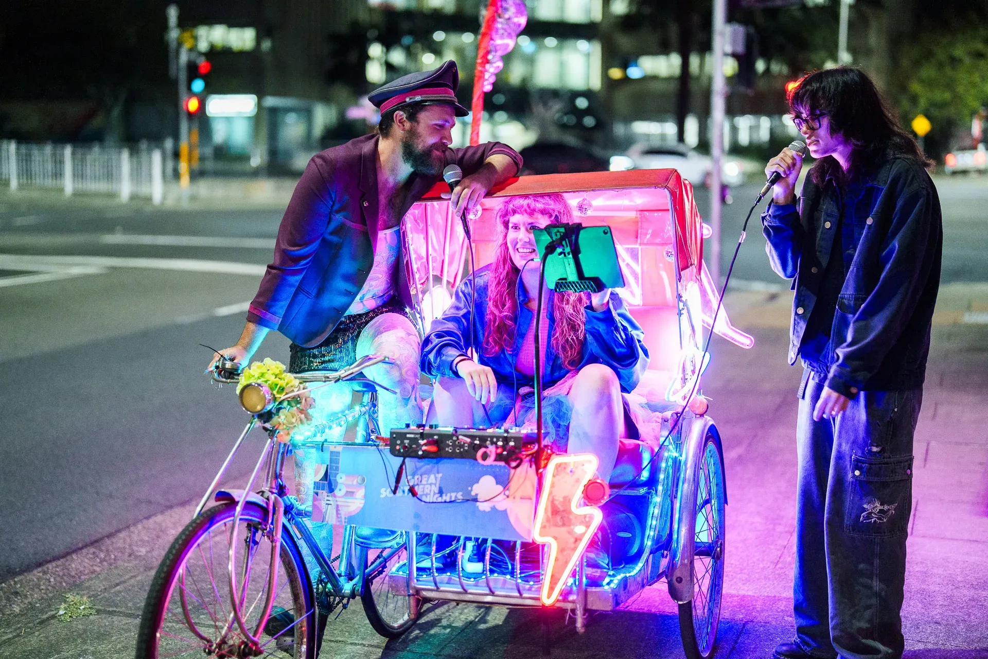 A person drives a brightly lit pedicab, while two other people sing into a microphone on a city street at night.