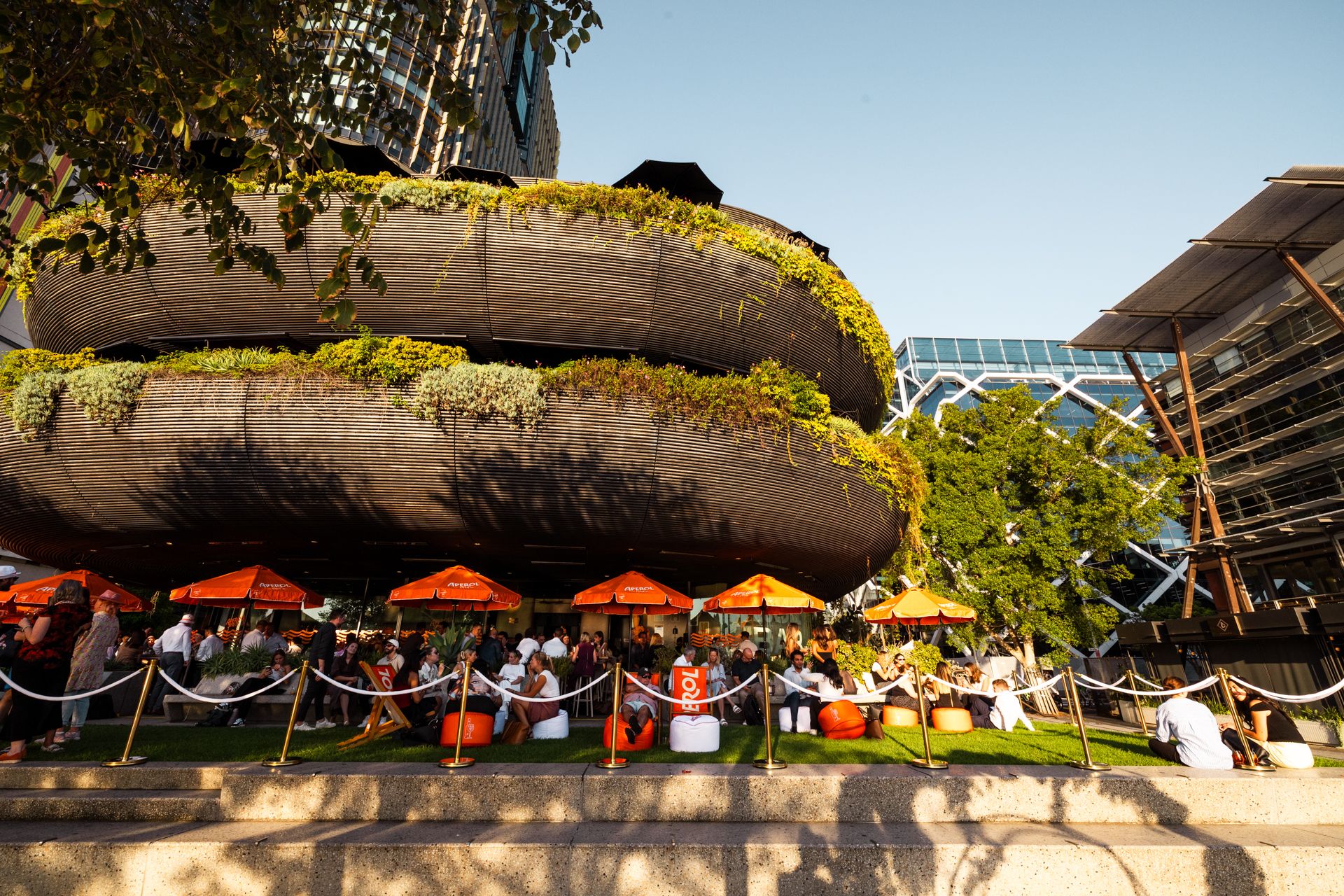 People relax on a grassy area with orange umbrellas outside a modern building adorned with greenery on a sunny day.