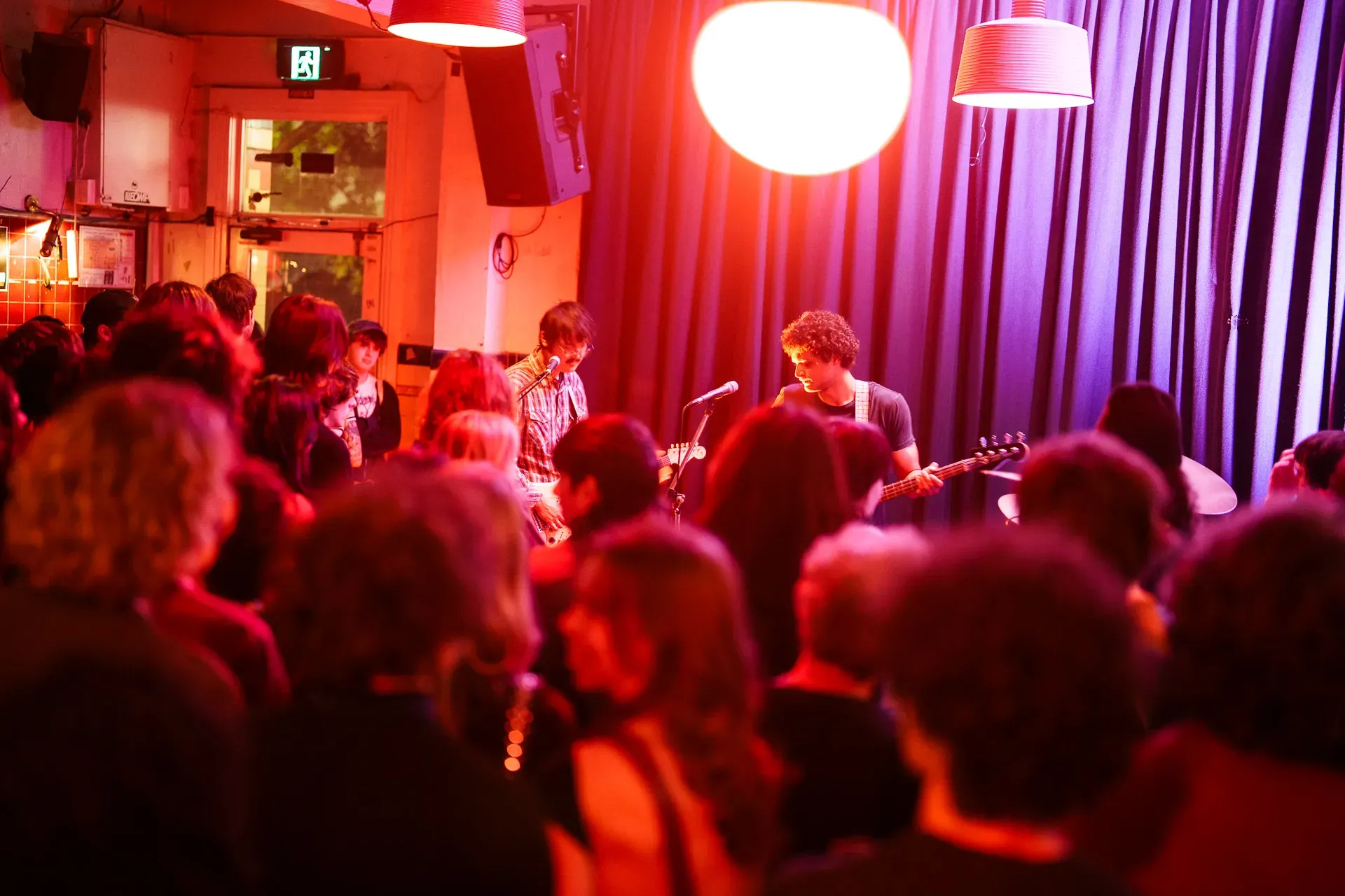 Musicians performing on stage in a dimly lit venue with a vibrant, red-hued crowd watching under hanging lamps.