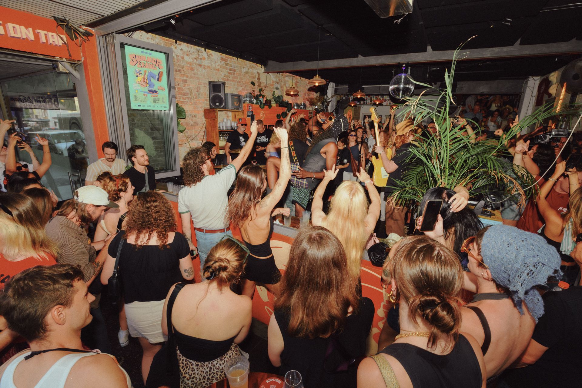 4 piece band with only 2 memebrs show in a sea of people surrounding both inside and outside the venue. A Big open window makes it accessible for people to view from the street which the shot shows. The tall dark and handsome lead singer dons a large sun hat with his hands in the air, whilst the red head guitarist with his back to the photo shreds a solo. Everyone in the crowd watch is ecstatic