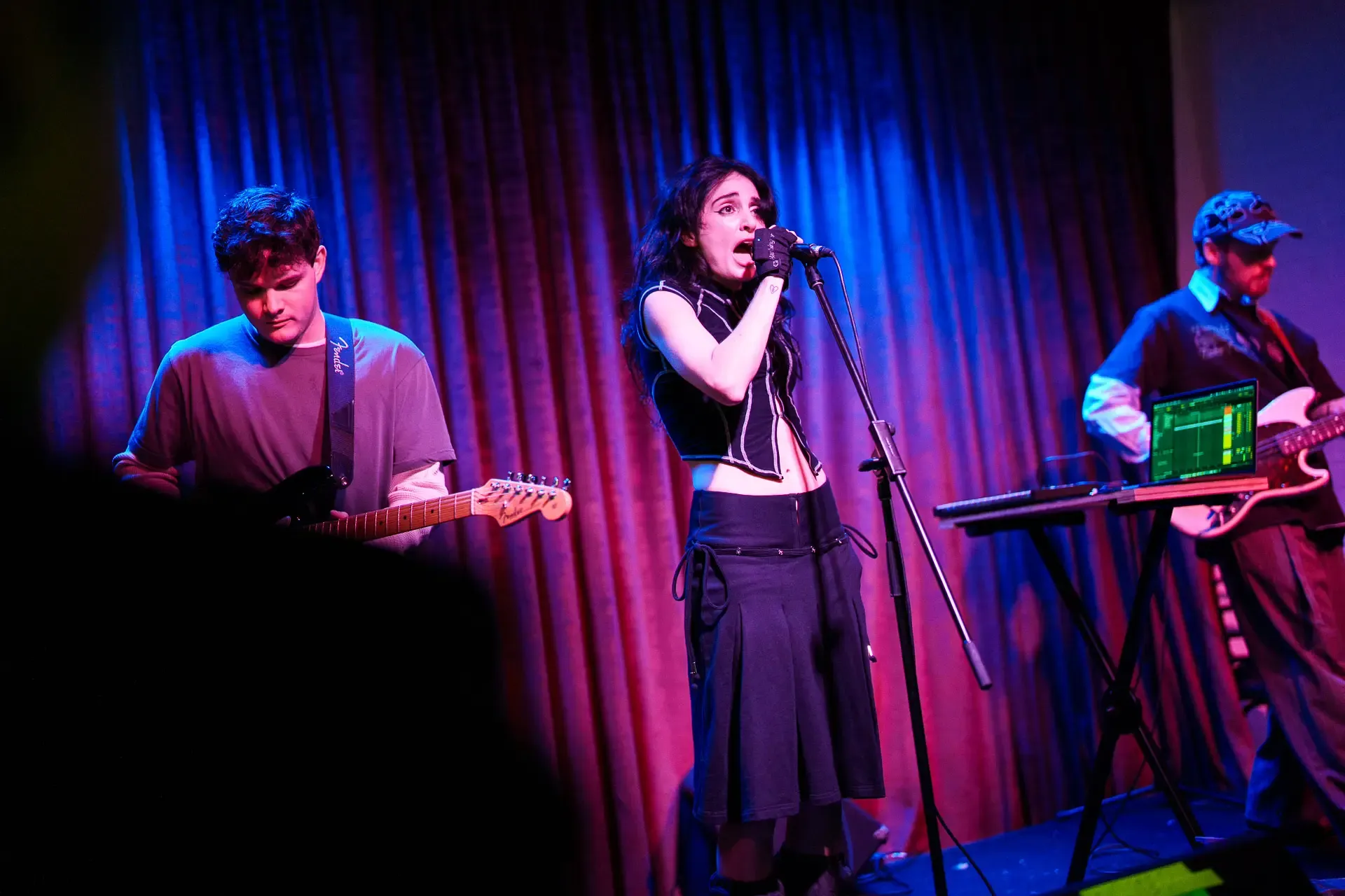 Band performing on stage under blue and purple lights. A woman sings into a microphone, flanked by guitarists, with a curtain backdrop.