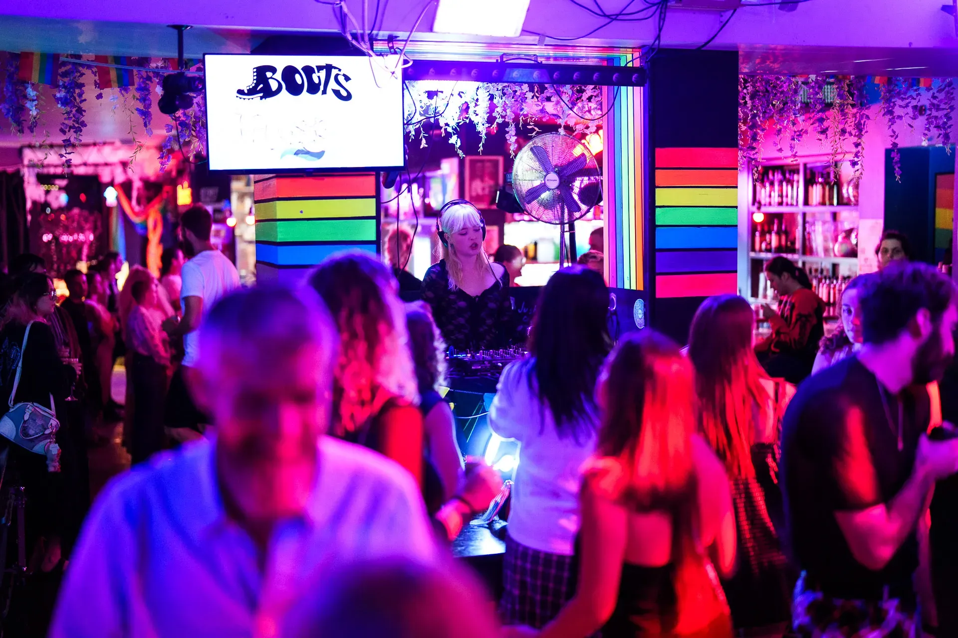 A DJ playing in a vibrant nightclub with rainbow decor, surrounded by a lively crowd under colorful lighting.
