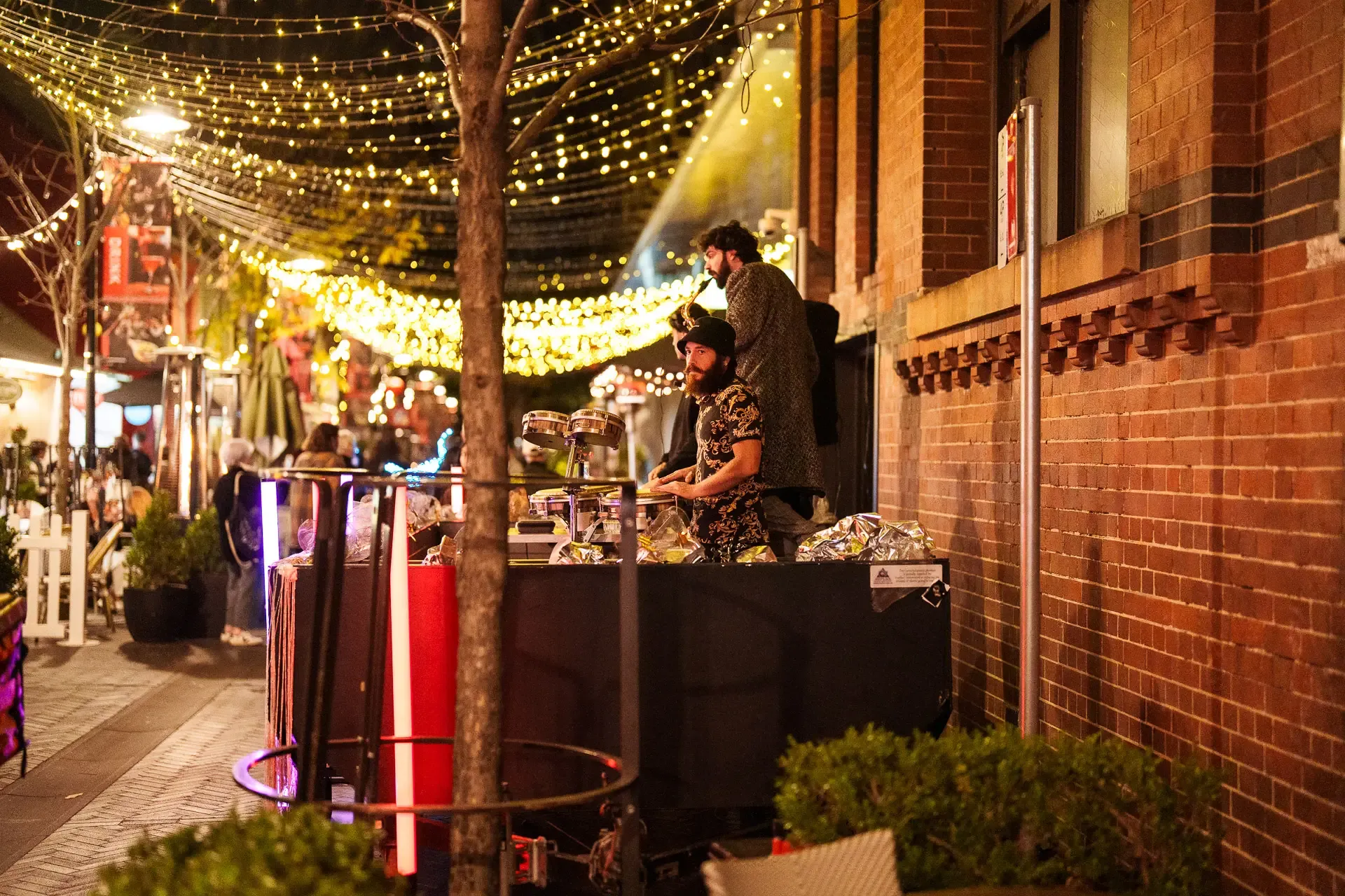 Street musicians perform under string lights on a lively, decorated urban street lined with pedestrians and shops at night.