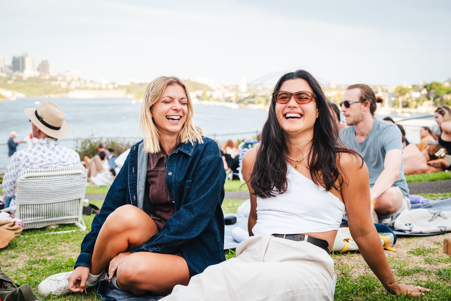 A couple young friends laugh and relax during Sunset Sessions, an open picnic concert series at Cockatoo Island, with the Sydney skyline visible in the background.