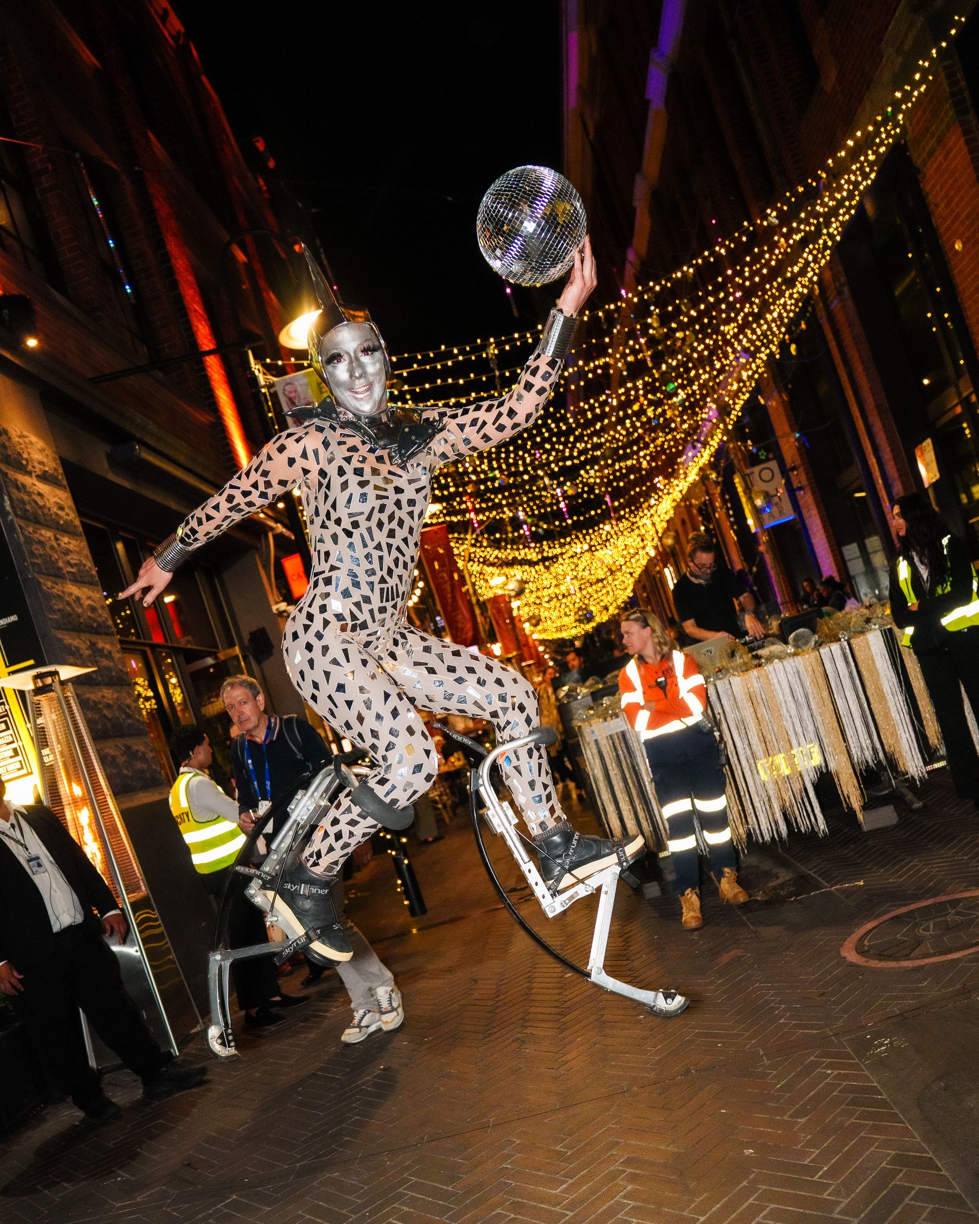 Street performer on jumping stilts in mirrored mosaic suit and silver face paint tossing a disco ball under canopy of string lights