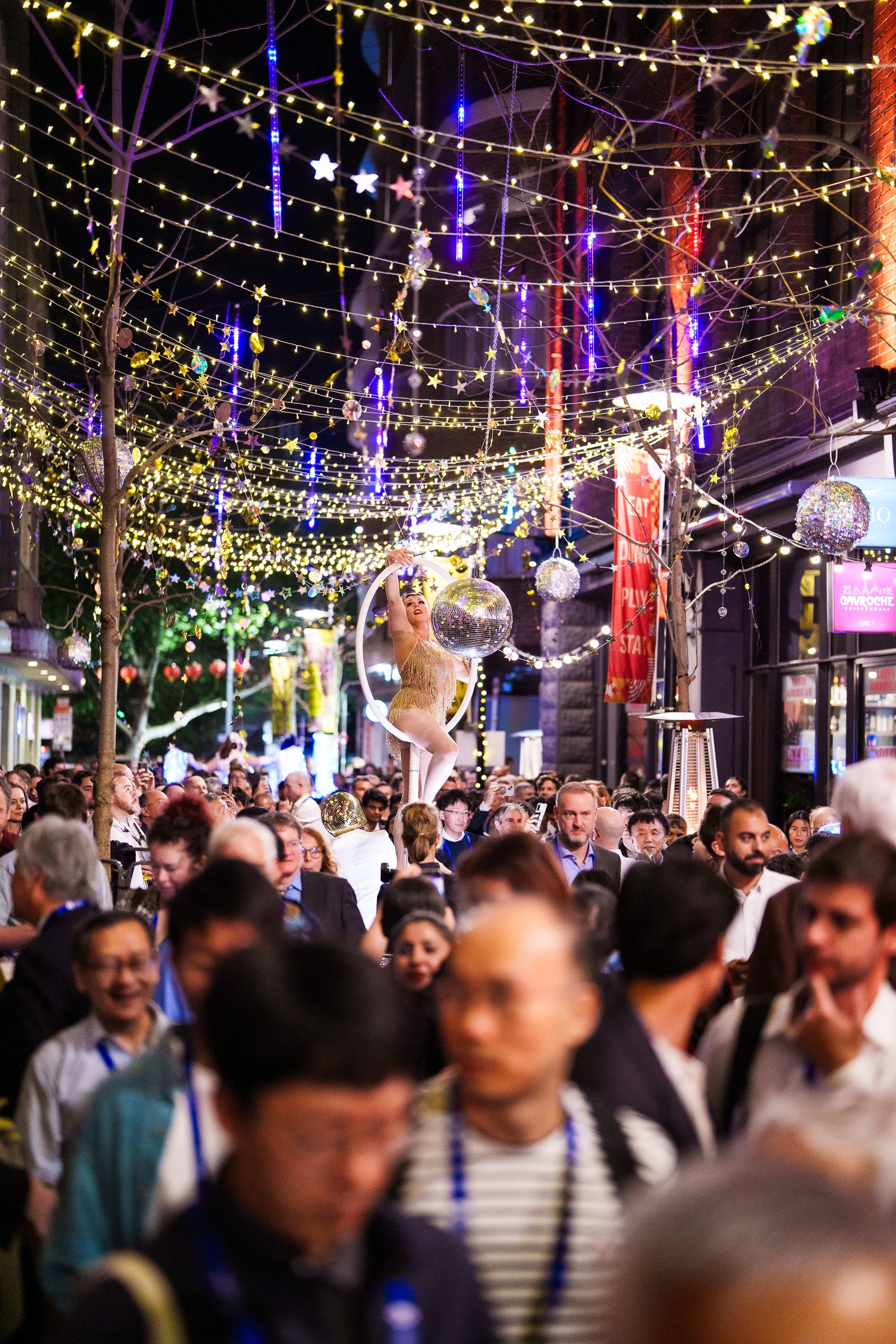 A crowded street at night with string lights and a performer in a hoop above the crowd.
