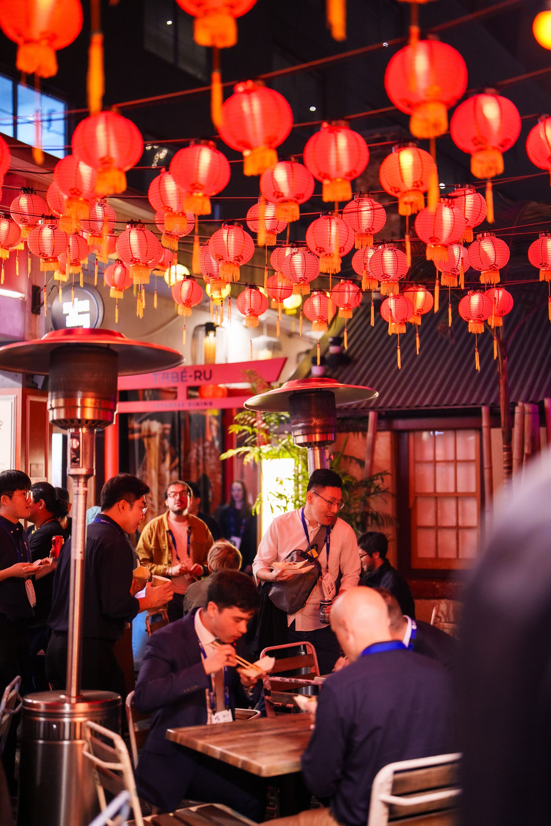 Crowded outdoor dining area under strings of glowing red paper lanterns, people eating and chatting at tables.