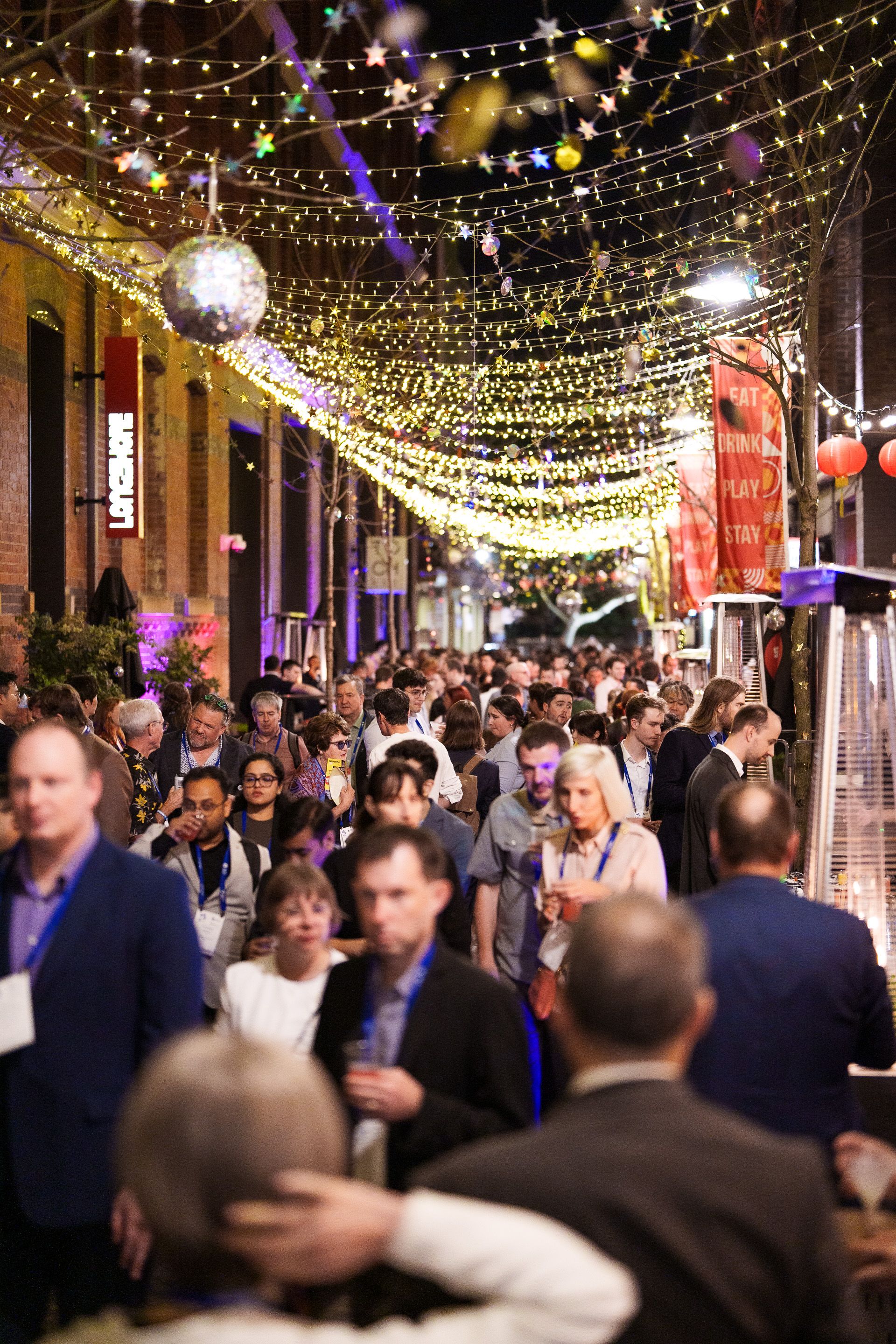 Crowded nighttime outdoor street scene with strings of fairy lights overhead and people socializing.