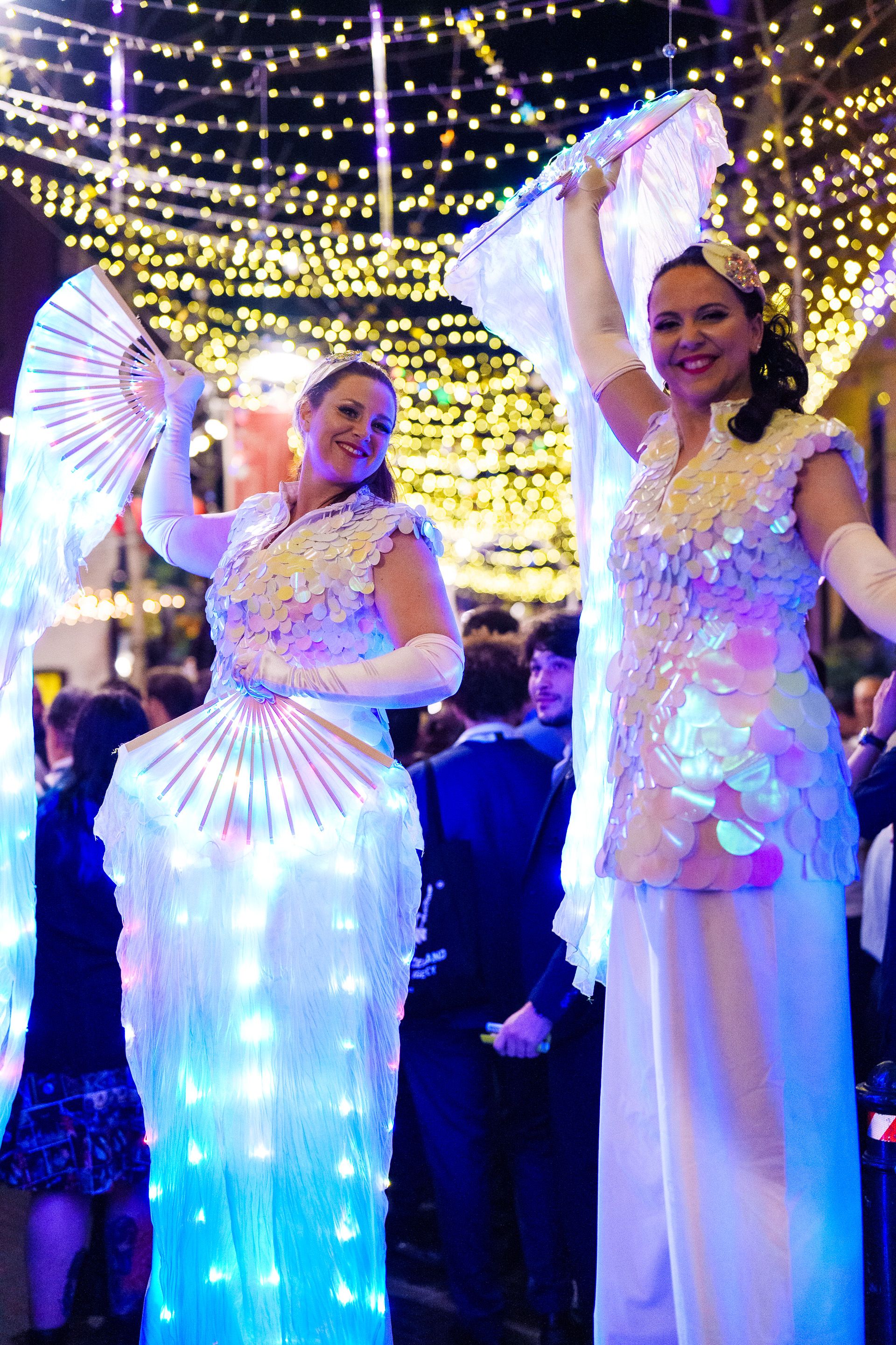 Two performers in iridescent sequin costumes with LED-lit flowing fans, smiling under a canopy of twinkling string lights.