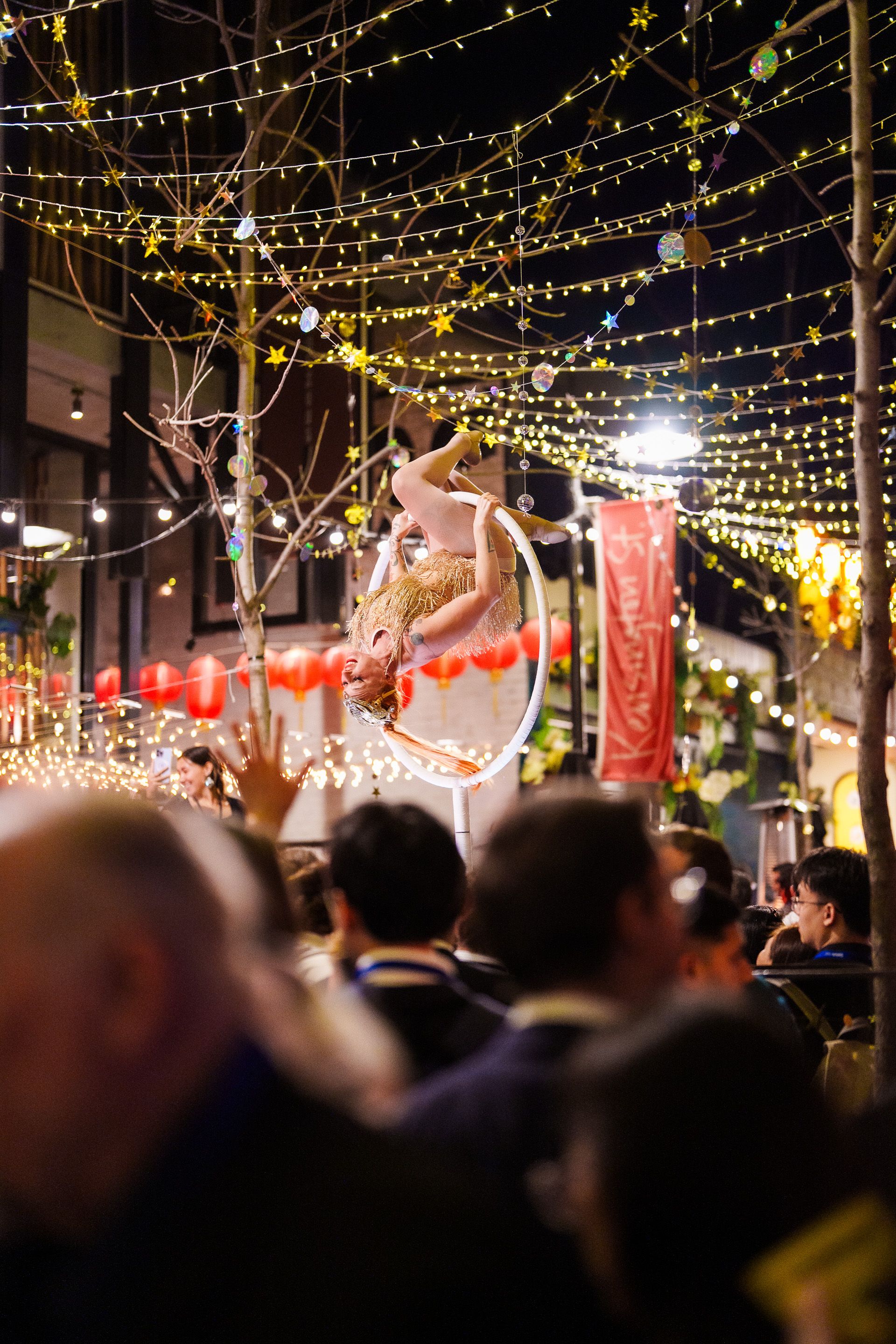 Aerial performer suspended upside-down in a hoop under twinkling string lights at a crowded nighttime street festival.