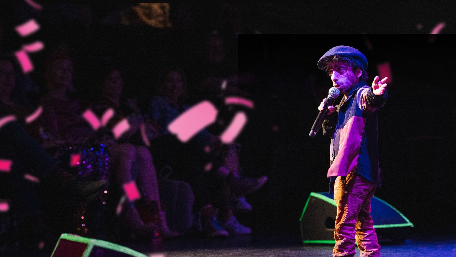 Performer in a cap on stage holds a microphone, extending one arm. Pink confetti falls around. Audience watches in the dimly lit background.