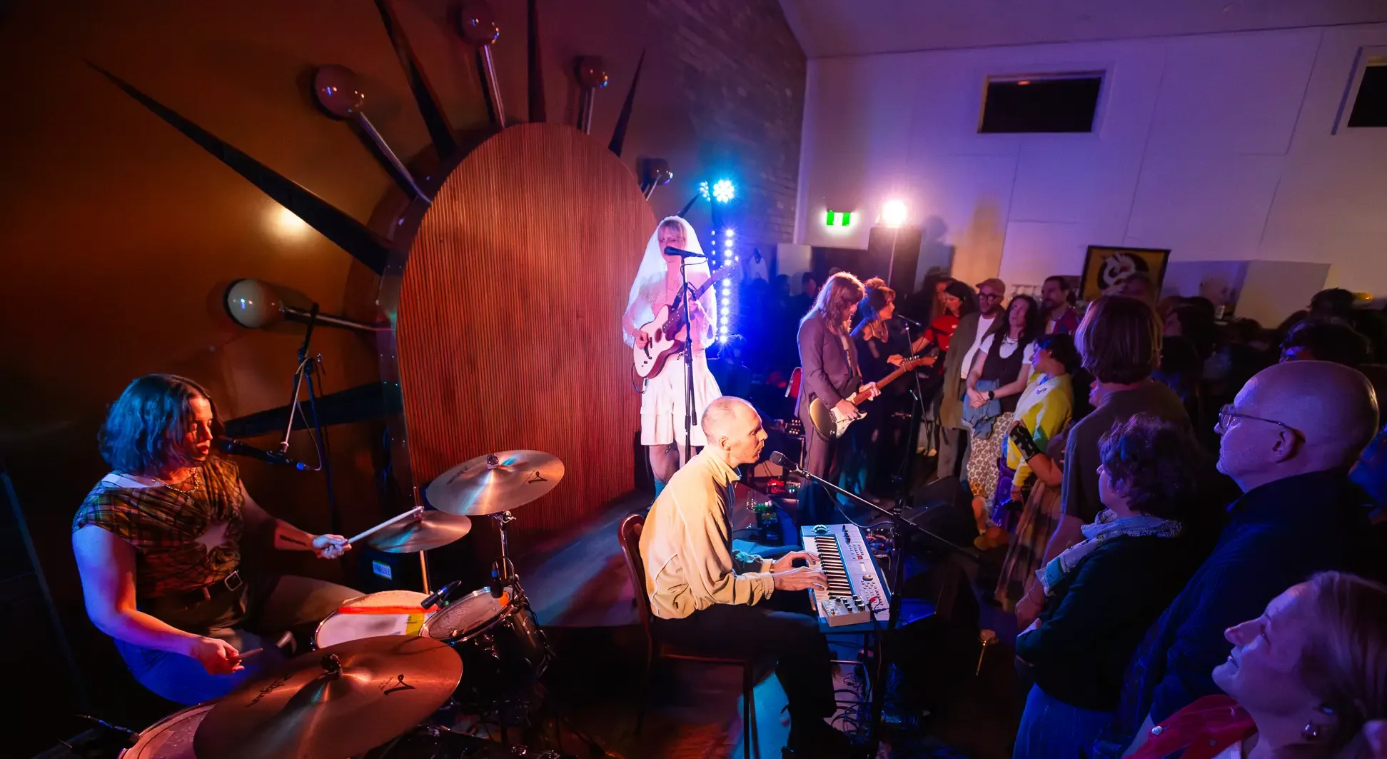 A band performs energetically on stage with a drummer, keyboardist, and guitarists in front of an engaged audience in a vibrant, colorful setting.