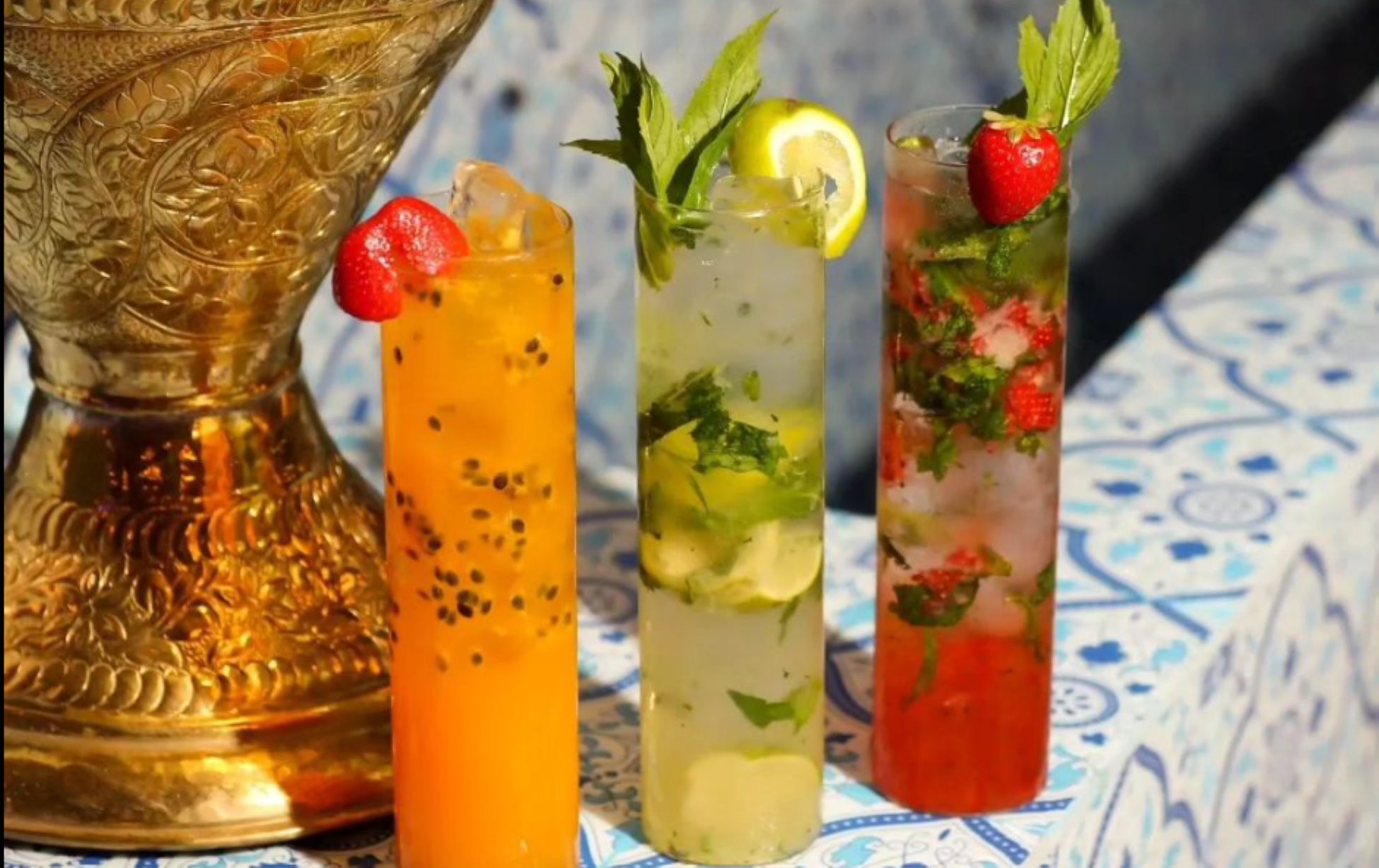 Three tall, colorful cocktails with fruit garnishes beside an ornate golden bowl on a patterned surface.