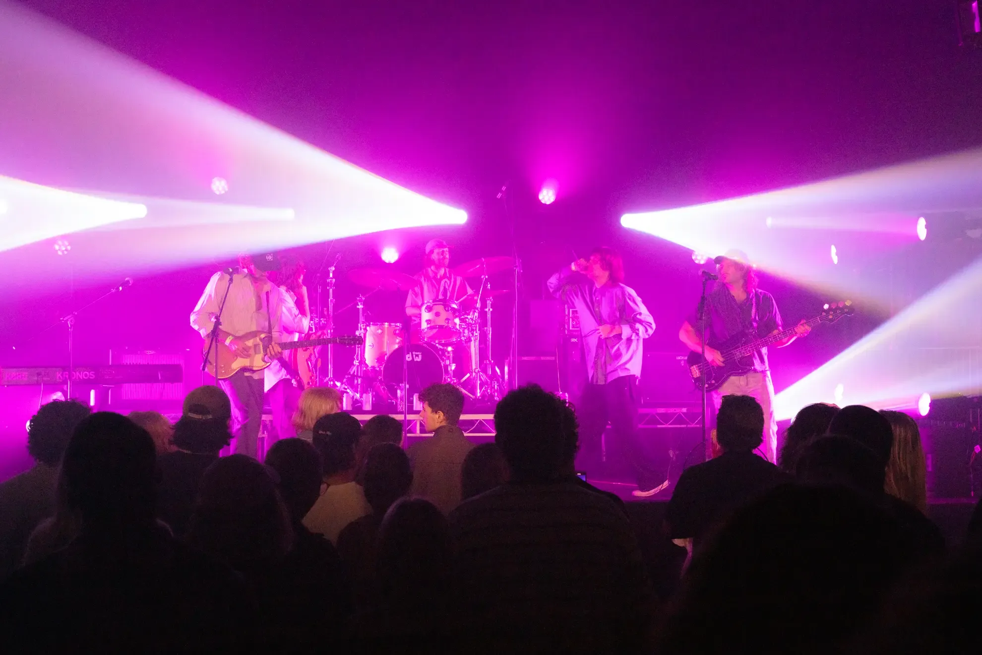 A band performs on stage with vibrant pink lighting, surrounded by an audience. Band members are playing guitar, drums, and singing.