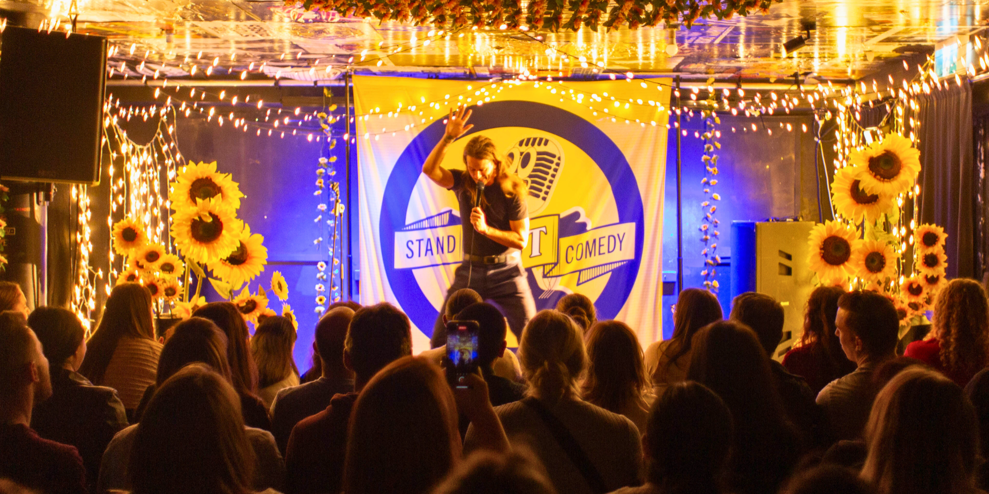 Comedian performing on stage with a microphone, surrounded by sunflowers and lights, in front of an engaged audience.