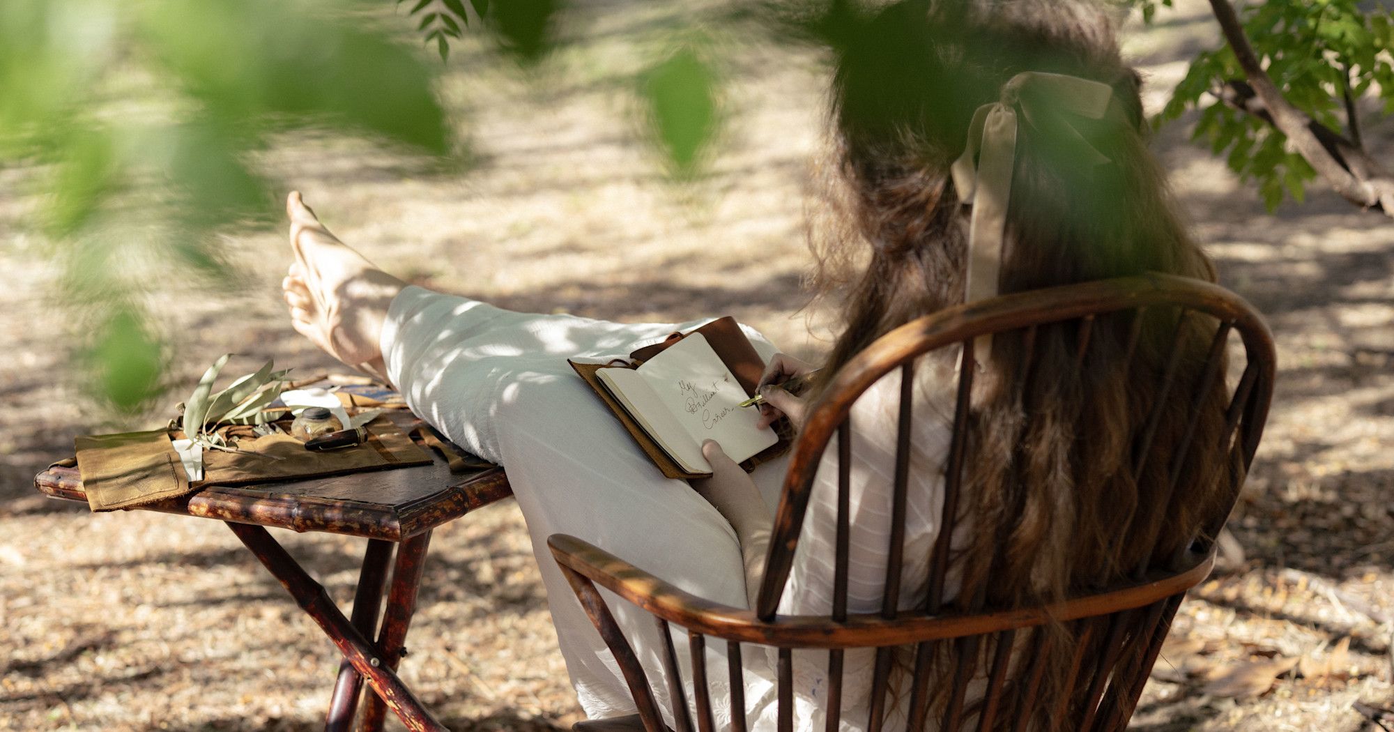 Person sitting in a wooden chair outdoors, writing in a notebook on their lap, with a small table nearby covered in various leaves and papers.
