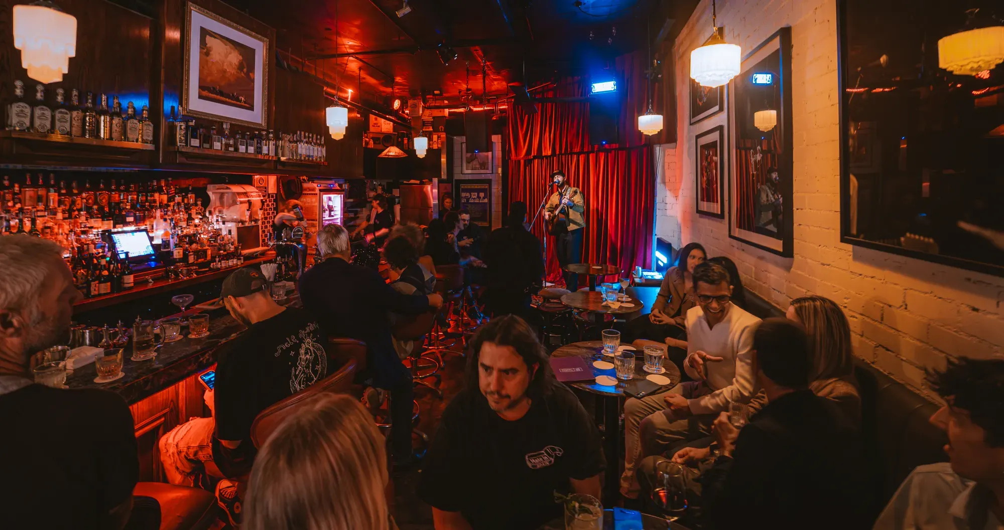 A cozy bar with dim lighting hosts a live music performance, attended by patrons enjoying drinks and conversations in an intimate setting.