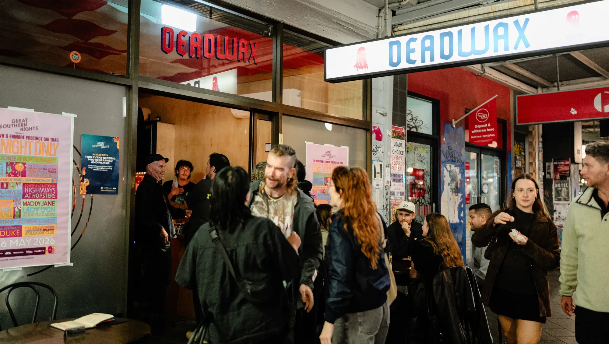People socializing outside a venue named "Deadwax" with posters and street art on the walls. Some are chatting while others enjoy food.