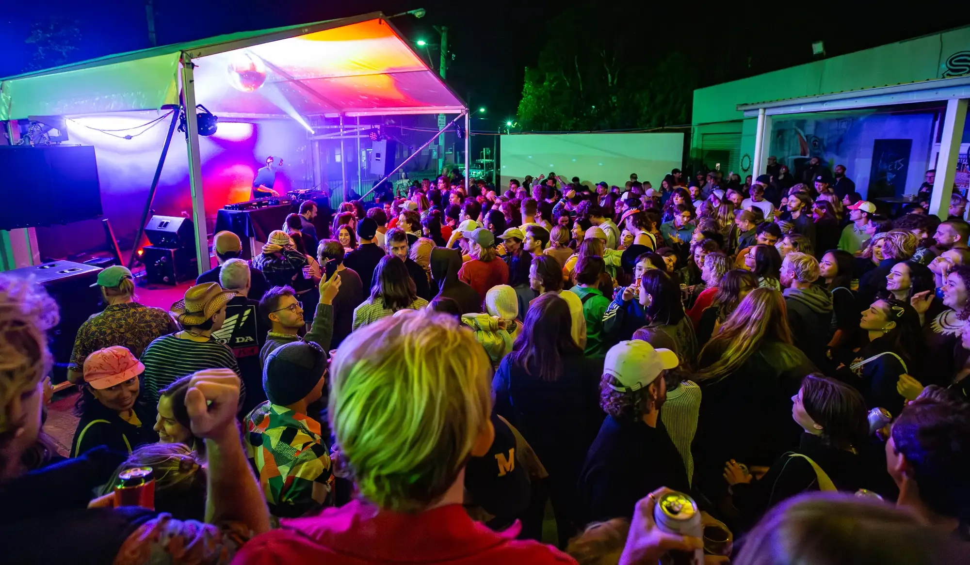 A lively crowd gathers at an outdoor night party, illuminated by colorful stage lights, with visible DJ equipment and vibrant atmosphere.