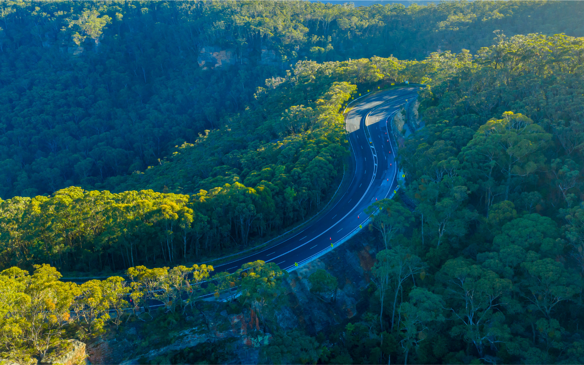 Aerial view of the Victoria Pass winding road through dense, sunlit forested hills, with shadows cast by the trees.
