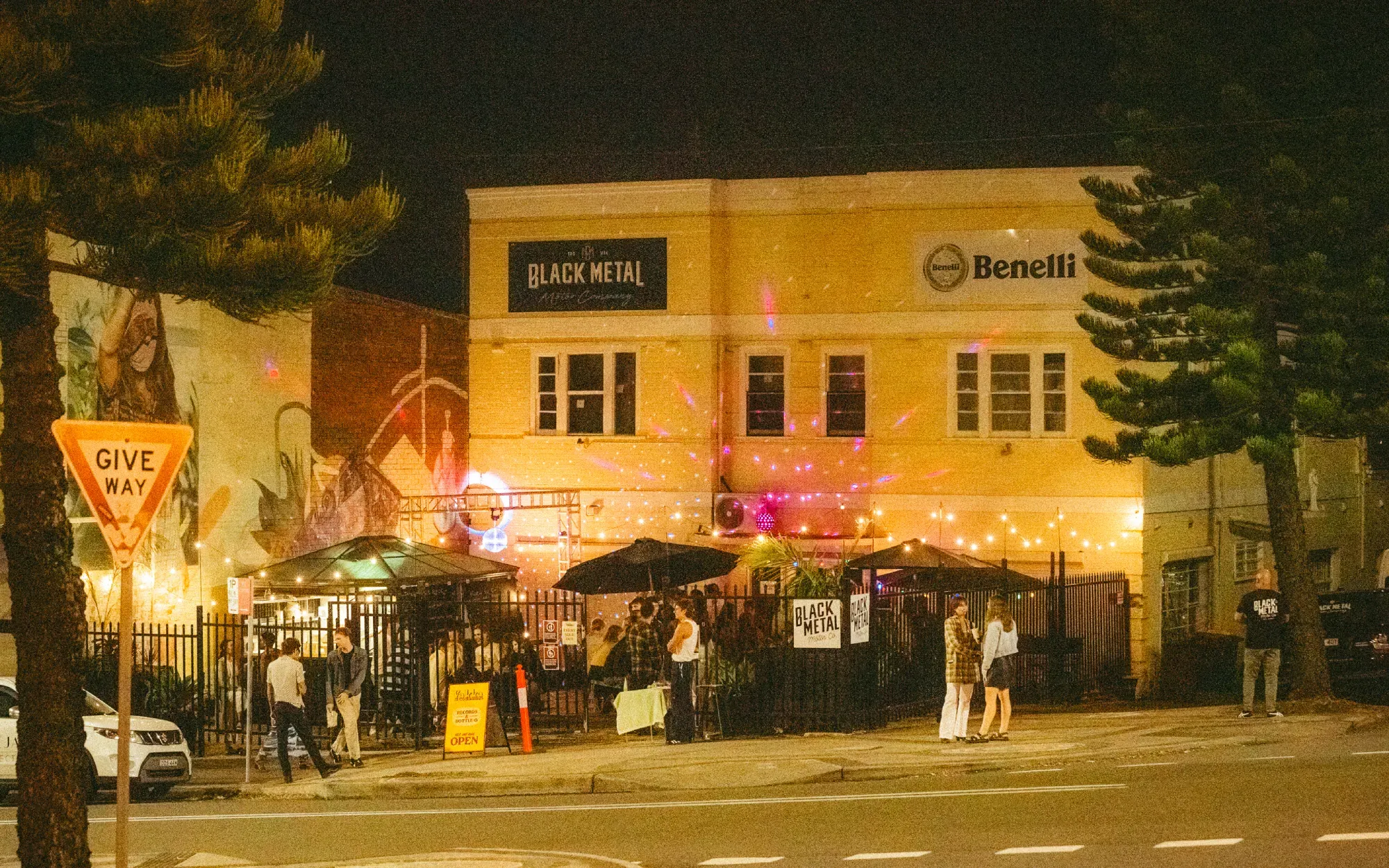 Nighttime street view of a lively venue with warm lights, outdoor seating, and people gathered, set against an urban backdrop.