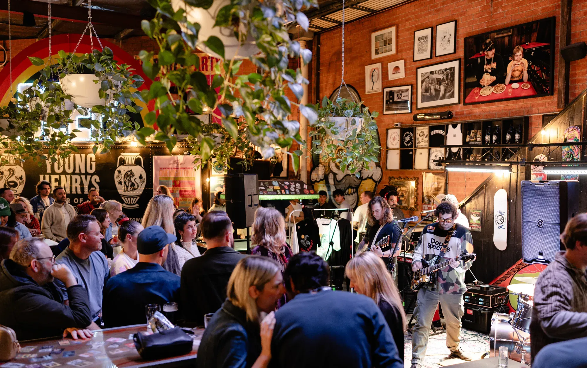 A lively indoor concert scene with a band playing and a crowd watching, surrounded by vibrant decor and hanging plants.