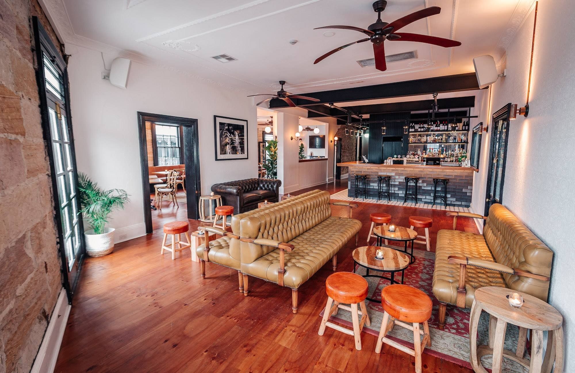 Stylish lounge with leather sofas, wooden stools, and a bar. Warm lighting, ceiling fans, and large windows create a cozy, inviting atmosphere.
