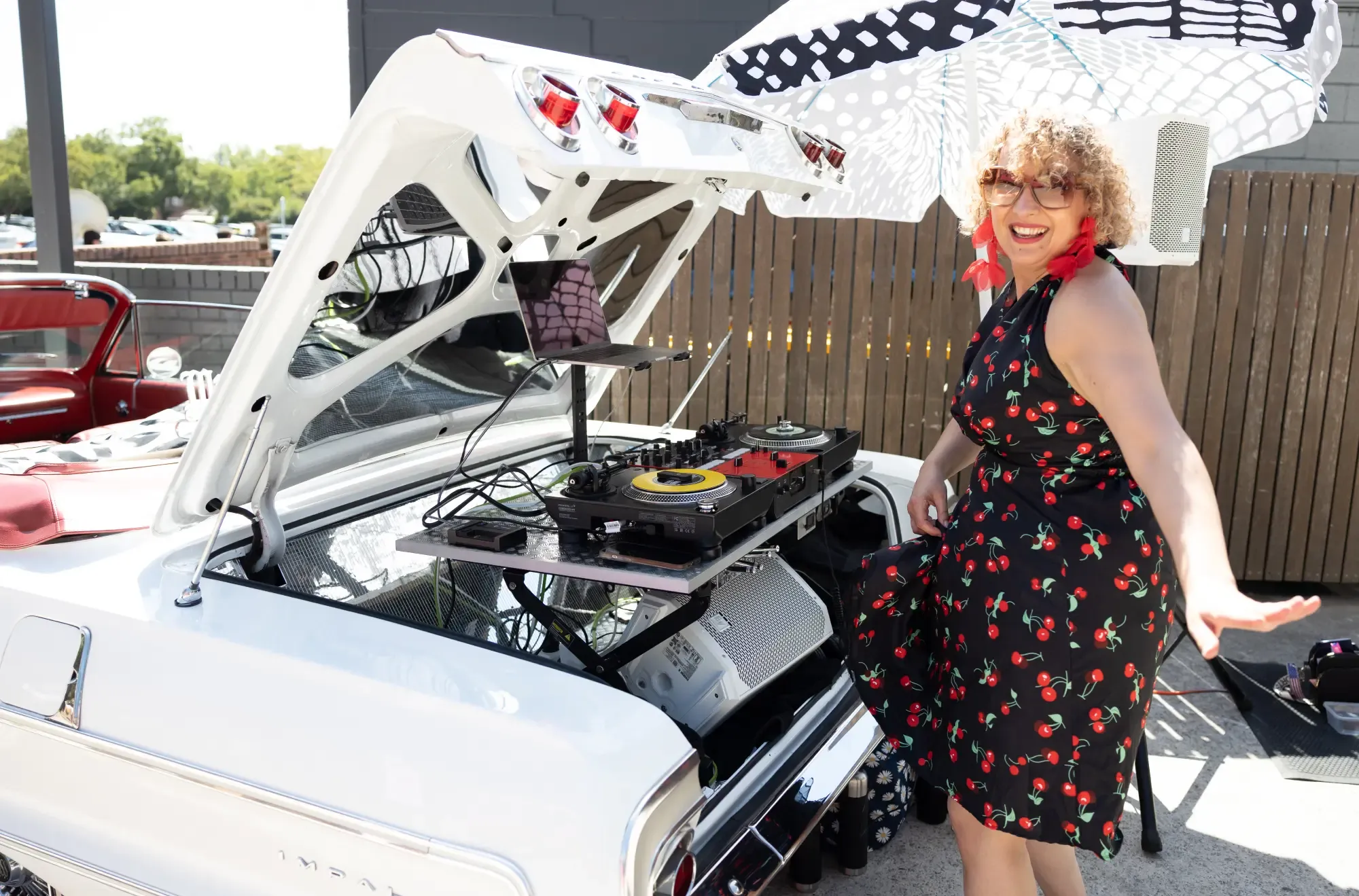 Woman in cherry dress DJing with turntables set up in a car trunk, holding a polka dot umbrella and smiling.