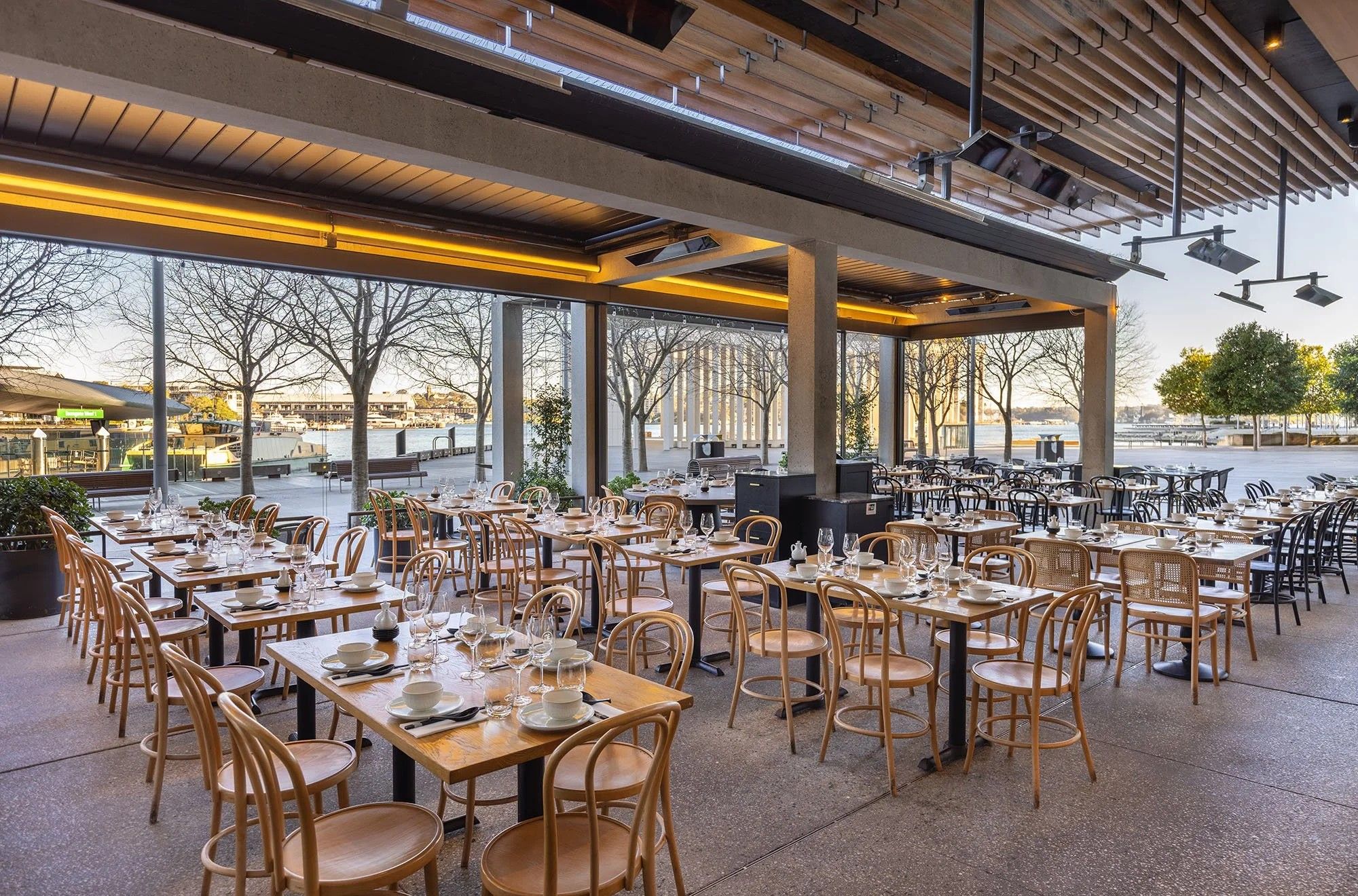 Spacious restaurant with wooden tables and chairs set for dining, large windows overlooking a waterfront view with trees and outdoor seating area.