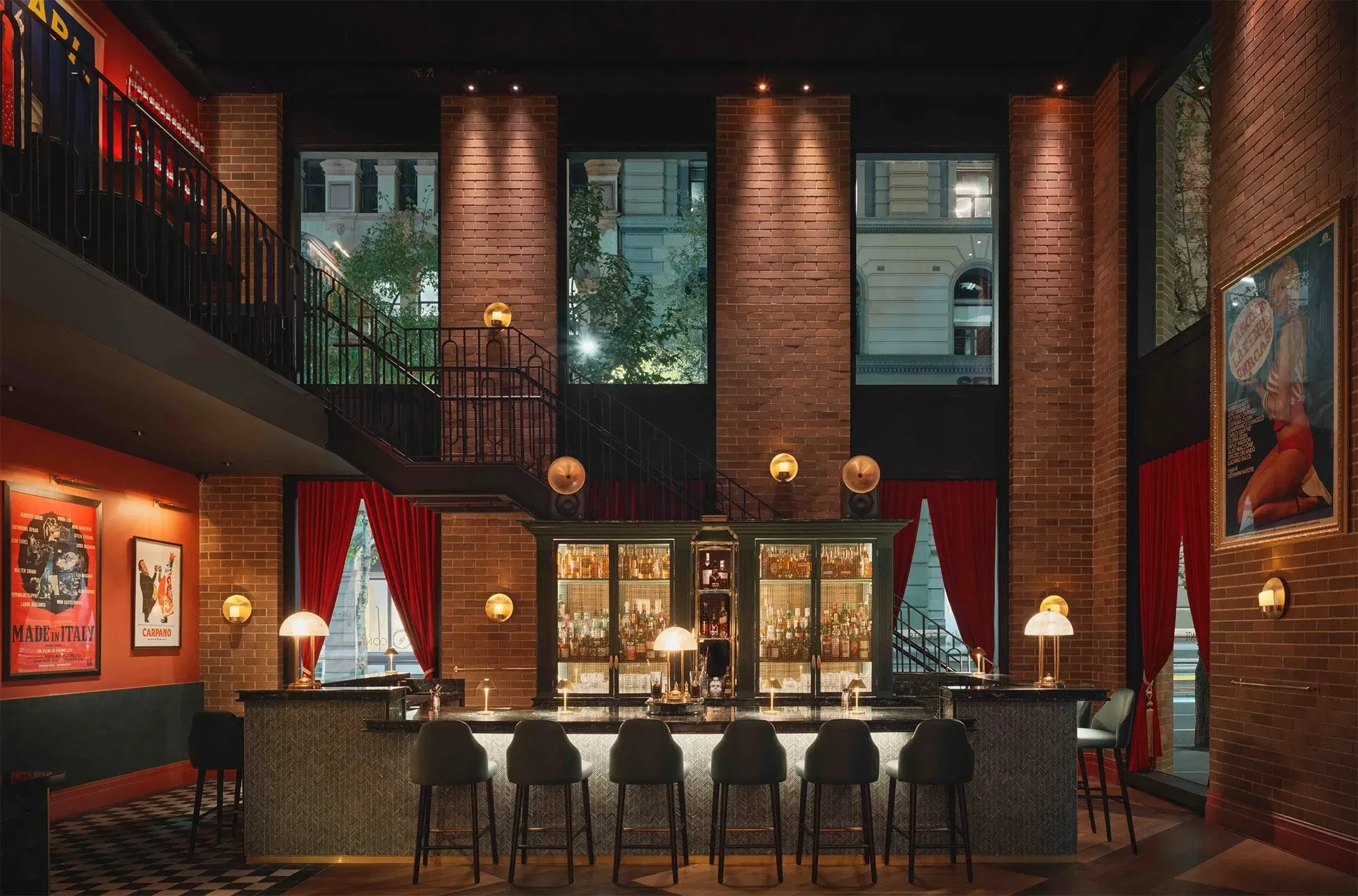 Chic bar with modern lighting, tall brick walls, and large windows. Red curtains and framed art enhance the stylish, upscale ambiance.