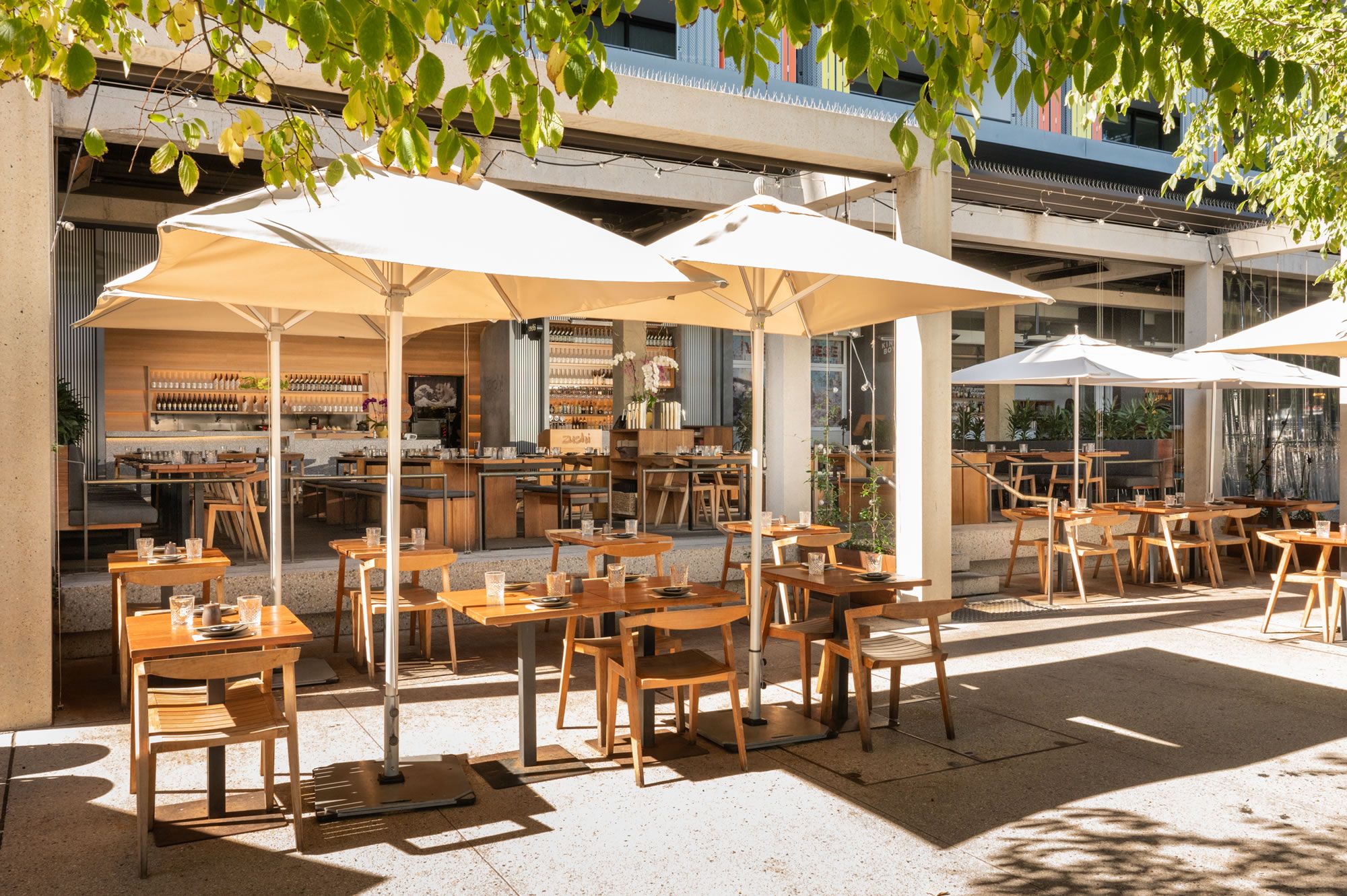 Outdoor cafe with wooden tables and chairs, shaded by large umbrellas. Sunlight filters through tree leaves, creating a relaxed, inviting atmosphere.