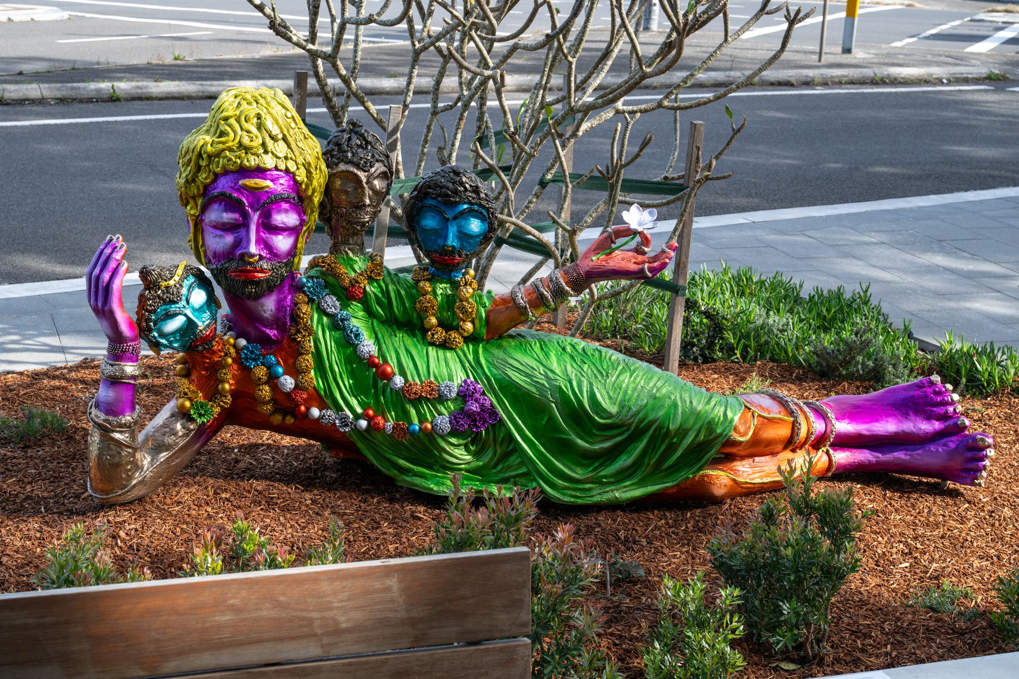Vibrant multi-headed reclining sculpture painted in purple, green, gold, and blue, adorned with colourful garlands and bracelets, holding a white flower, installed in a landscaped public space.